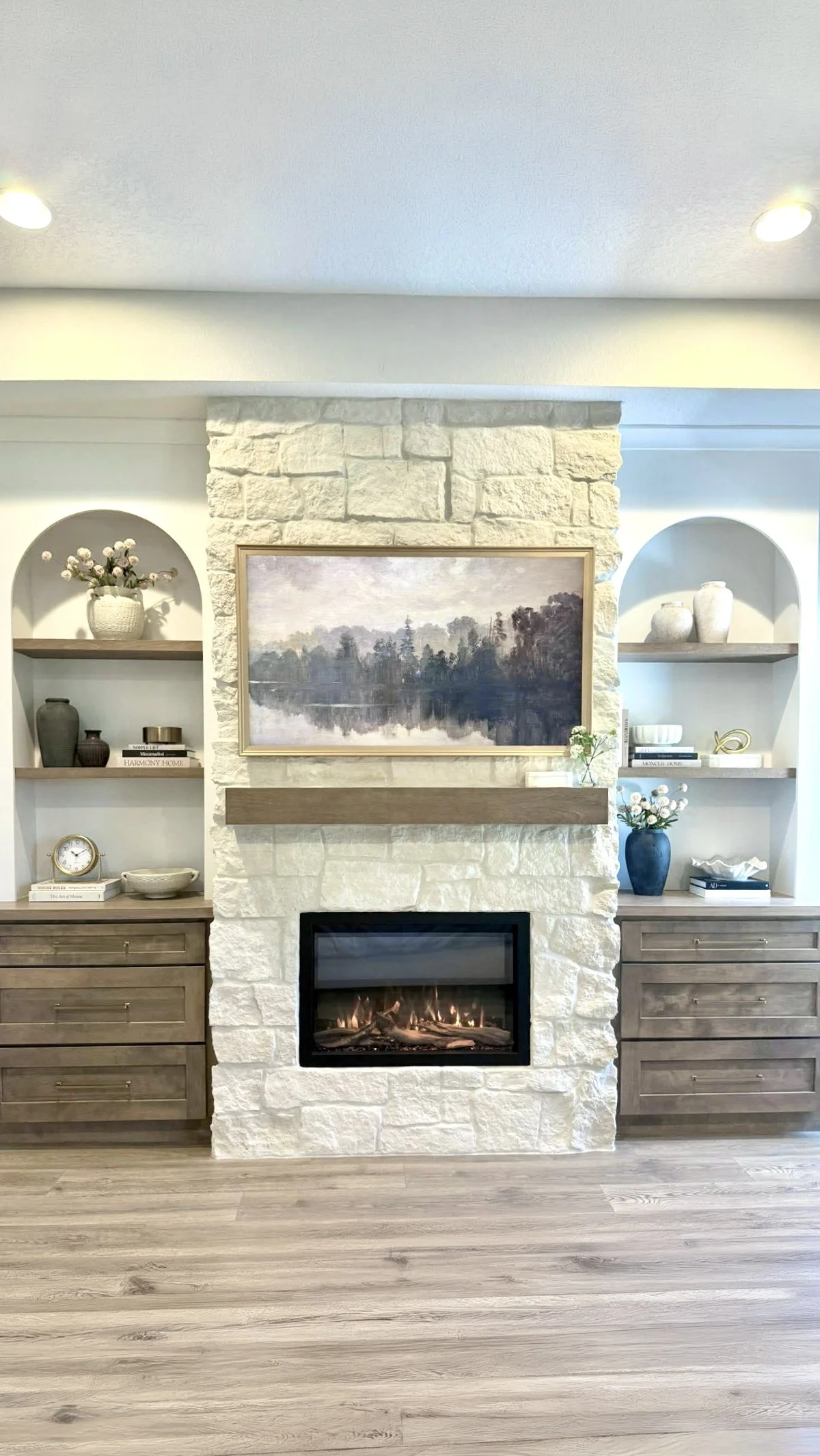 Living room with a white stone fireplace, framed landscape painting, built-in shelves with decorative vases, books, and a clock, and wood flooring.