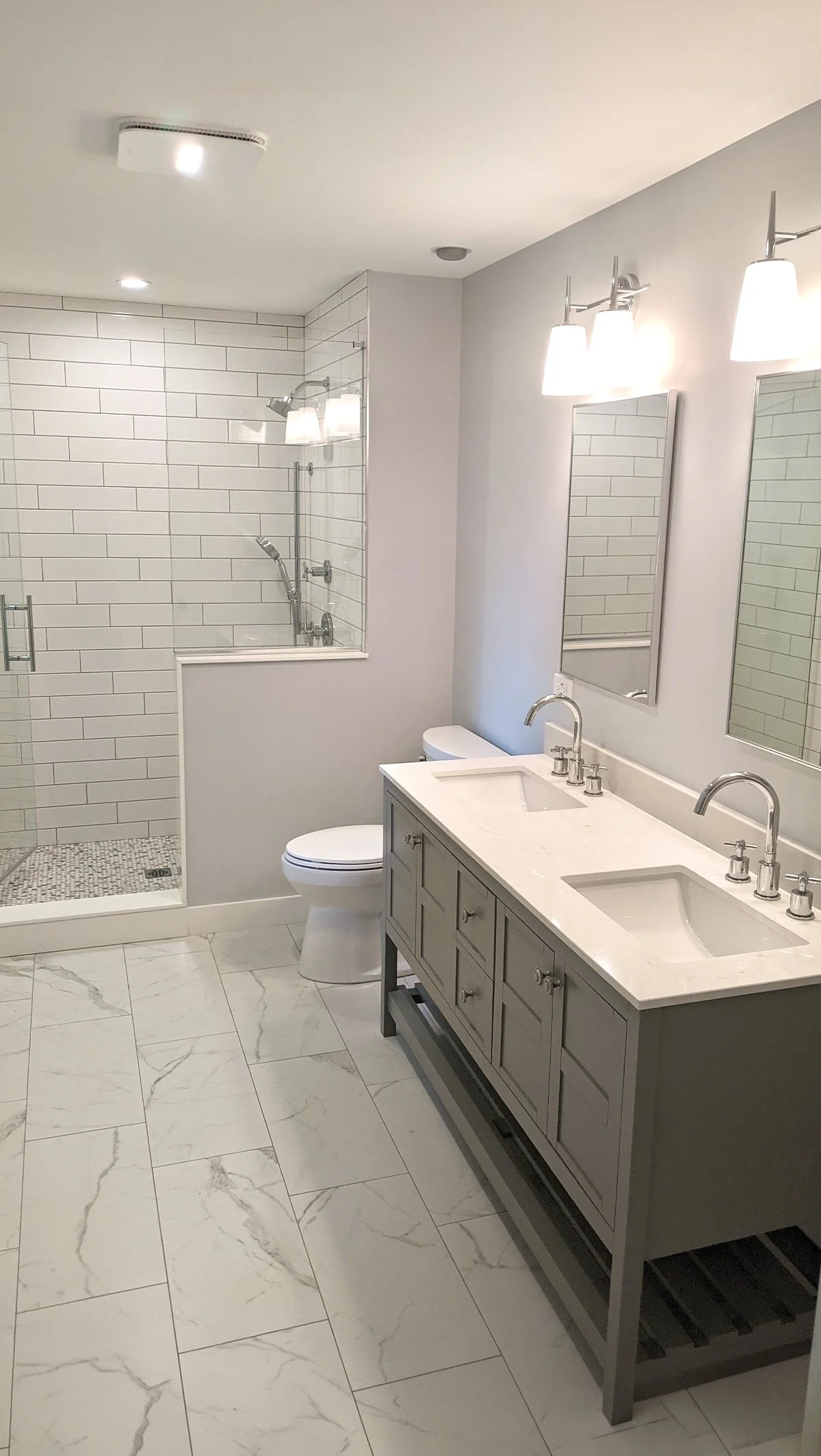 Modern bathroom with dual sinks, a light gray vanity, large mirrors, a toilet, and a walk-in shower with a white subway tile wall and a pebble tile floor inside.