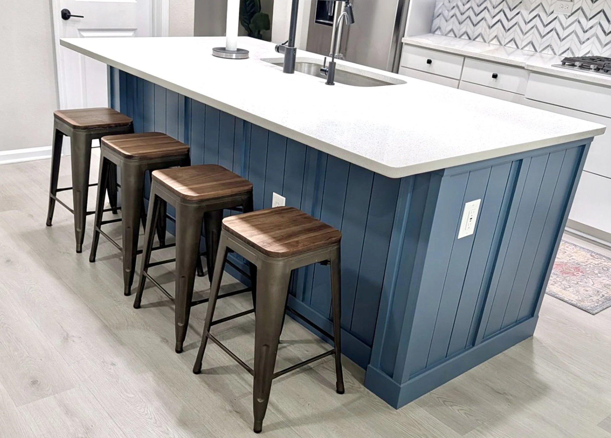 Kitchen island with blue paneling, white countertop, three wooden bar stools, stainless steel sink and faucet, gray backsplash, and appliances.