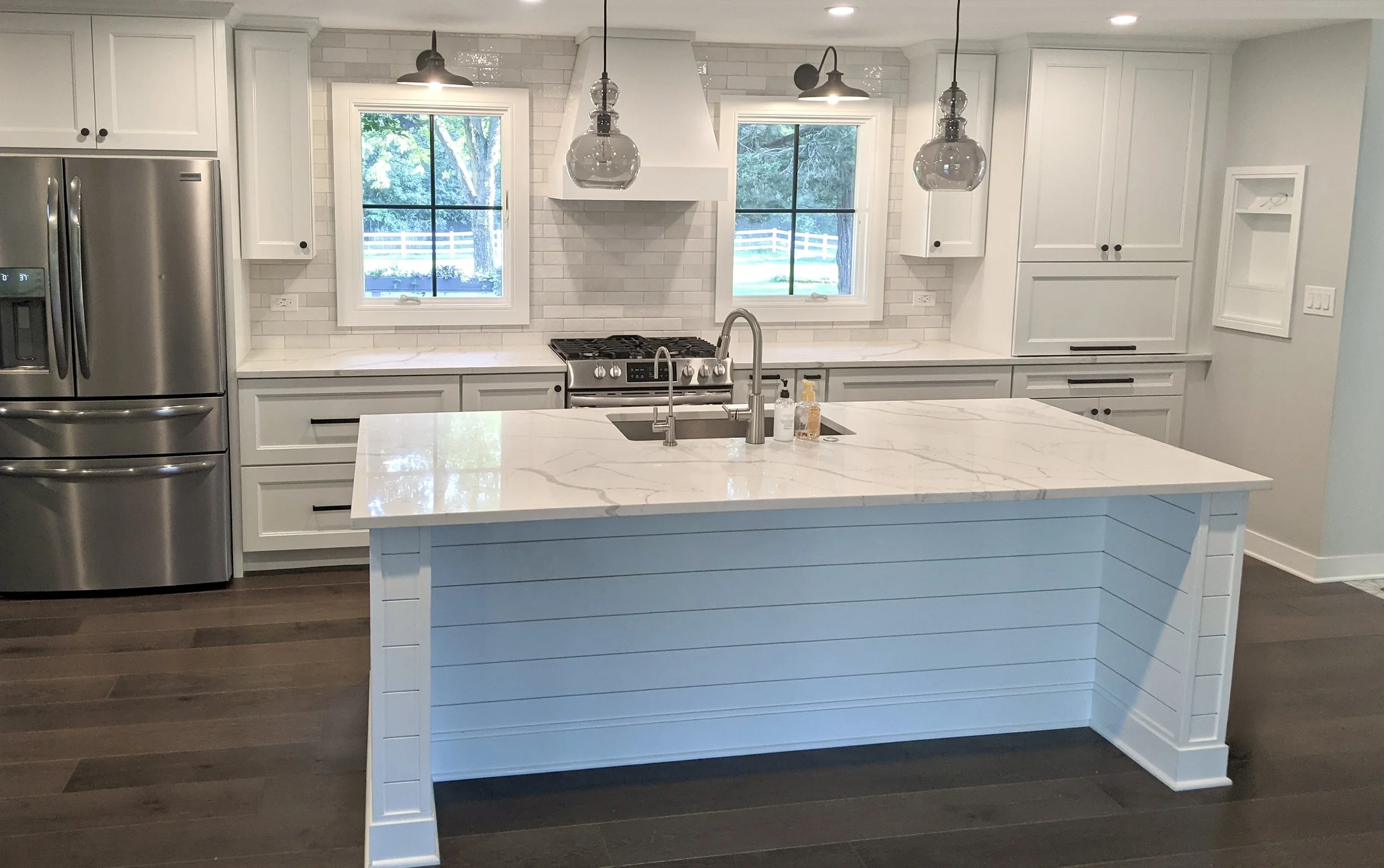 Modern kitchen with white cabinets, a white island with a marbled countertop, stainless steel refrigerator, and a gas stove under two large windows showing an outdoor scene with trees and a white fence.