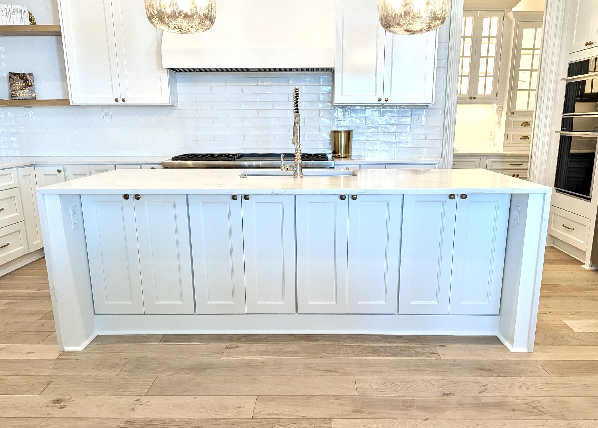A modern kitchen with white cabinets, a white kitchen island with a marble countertop, a gold-colored cup, a stainless steel sink with a flexible spring faucet, white subway tile backsplash, and hardwood flooring.