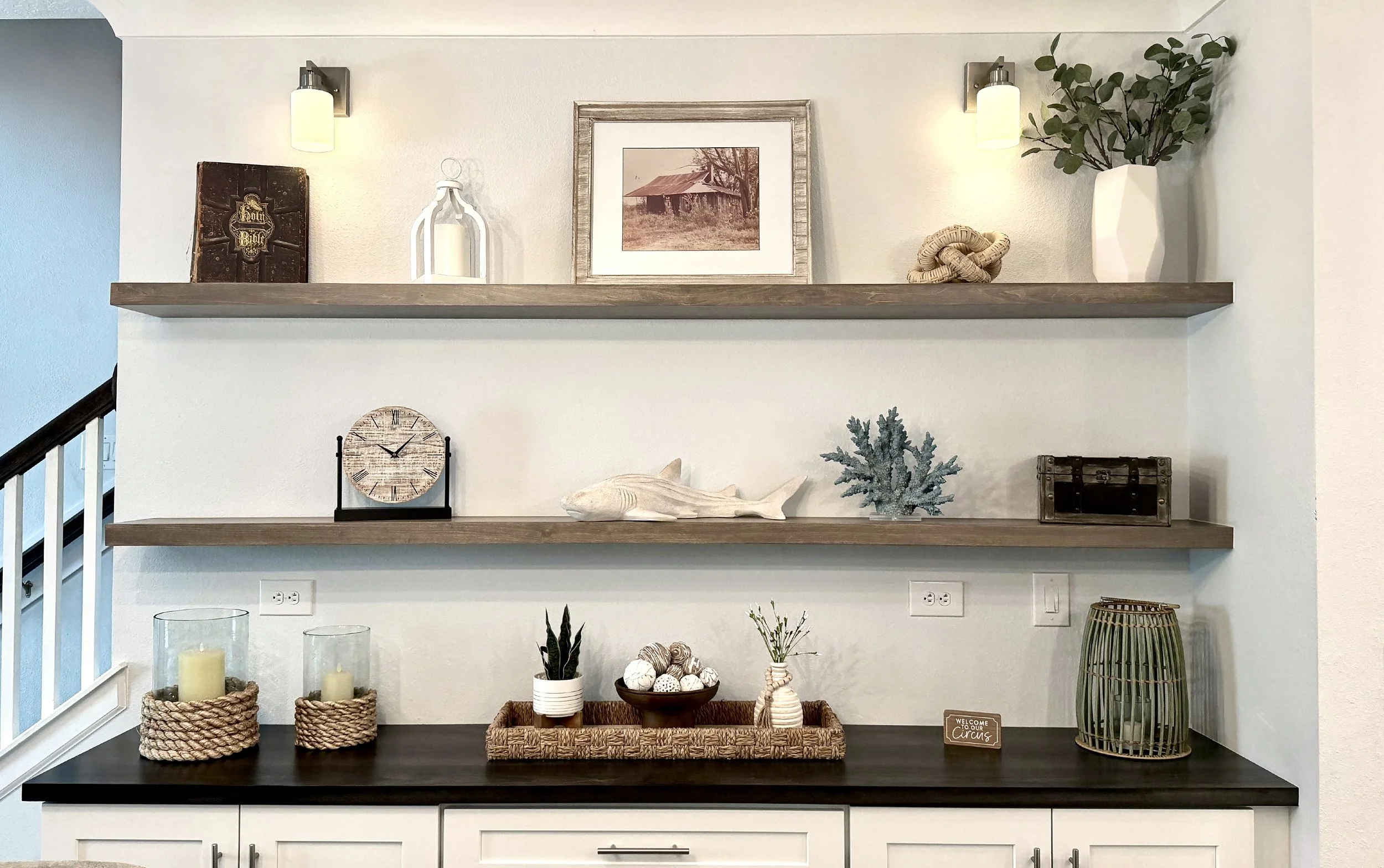 Decorative wooden shelves with farmhouse-style décor including a framed picture of an old barn, a white lantern, a ceramic vase with greenery, a woven knot, and a large white vase with leafy branches, mounted on a light-colored wall with wall-mounted lights.