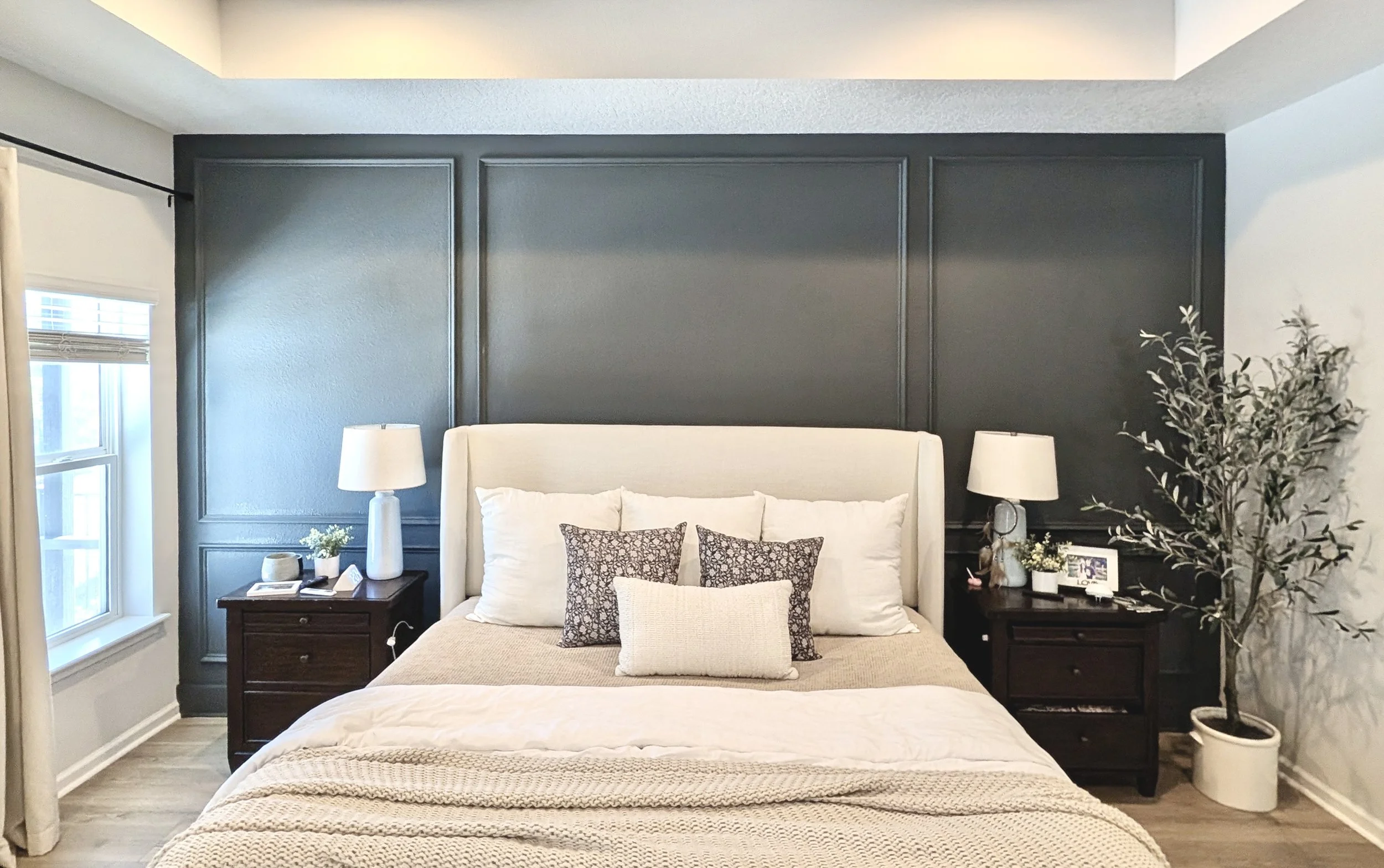 A bedroom with a bed, nightstands, and lamps on each side, large windows, a dark feature wall, and decorative pillows.