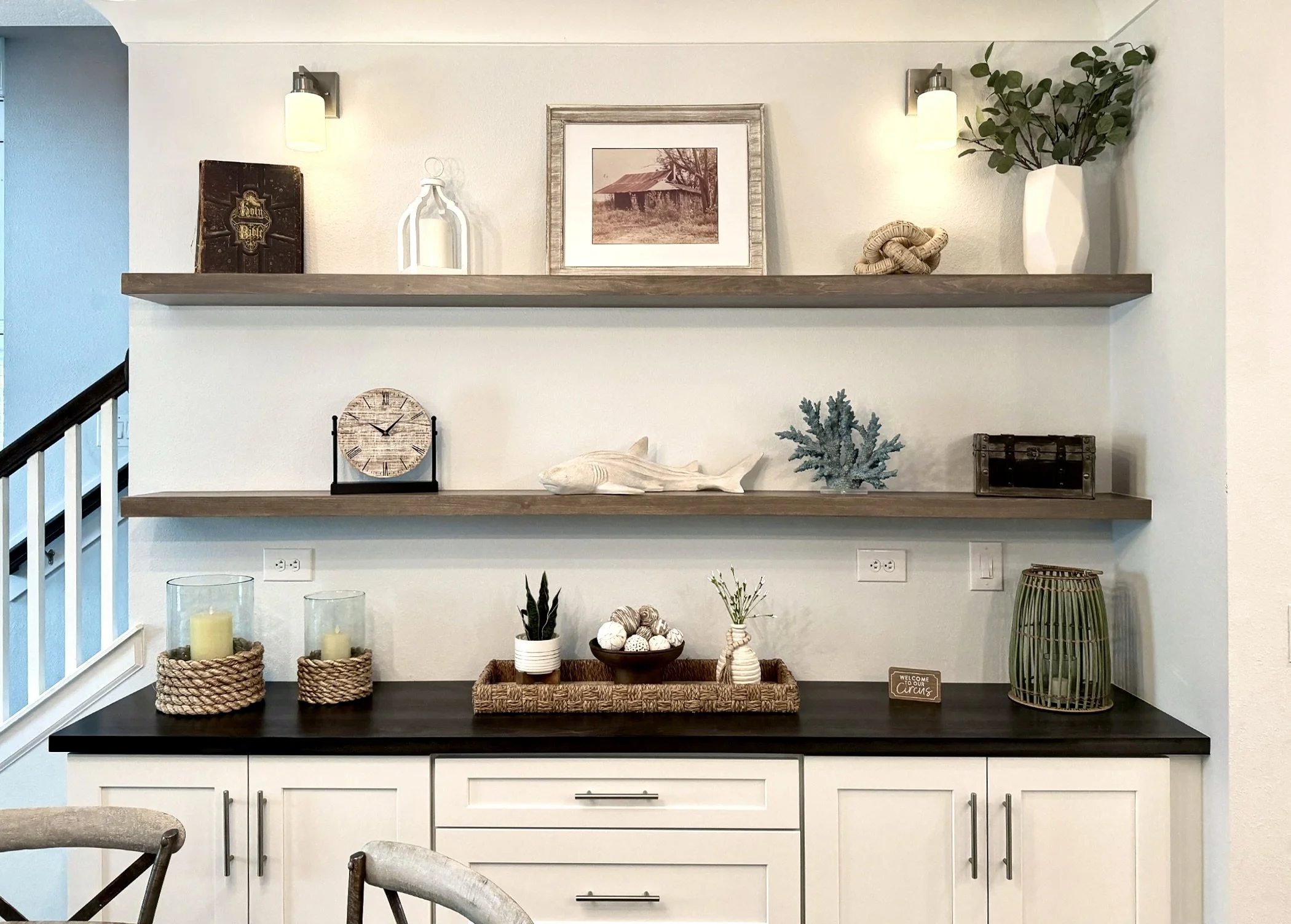 Decorative farmhouse style living room wall with two floating wood shelves holding various decor items, including framed artwork, vases, plants, and a book, with a section of white cabinetry below.