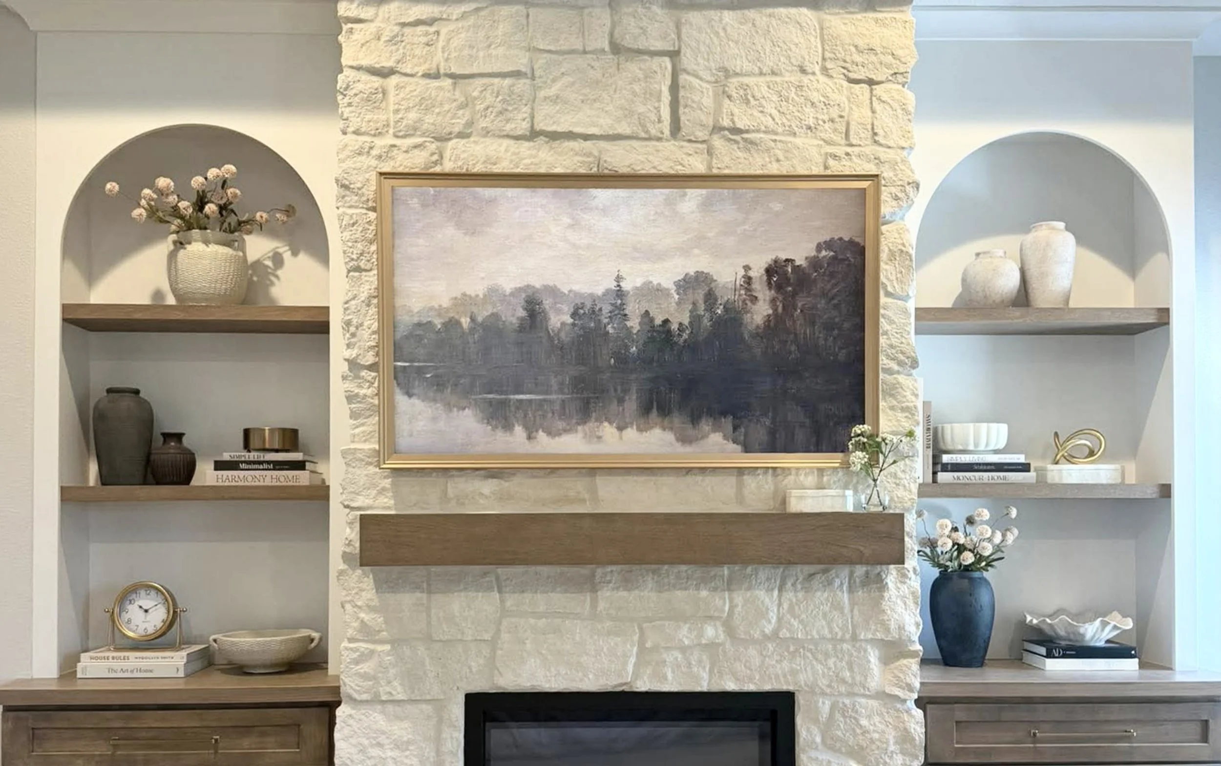 Living room with a white stone fireplace, framed landscape painting of a lake and trees, and built-in wooden shelves with decorative items including vases, books, and flowers.