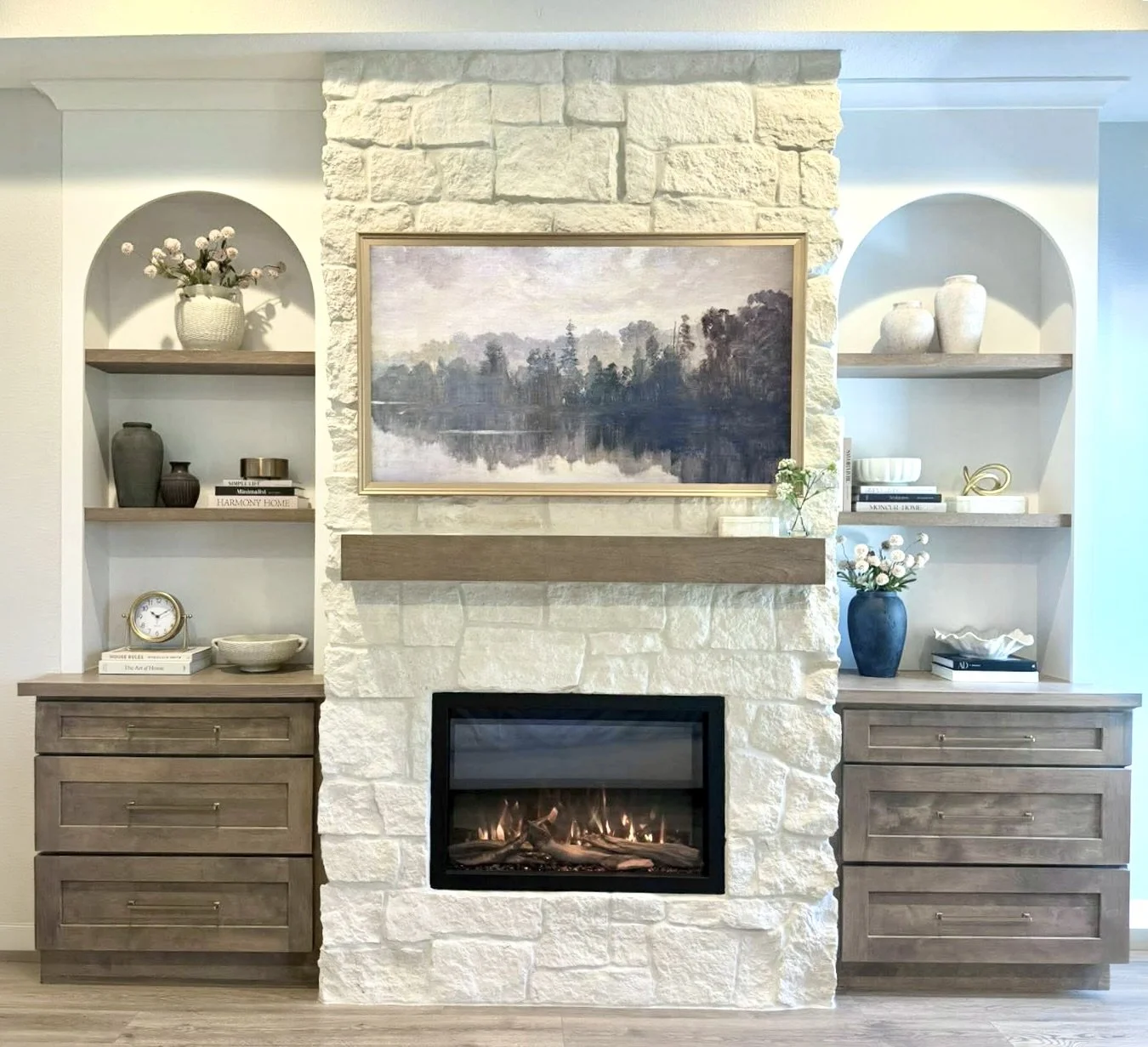 Living room fireplace with a digital fire, built into a white stone wall. Above the fireplace, a landscape painting of a lake and trees hangs. Flanking the fireplace are light wood cabinets with open shelves displaying decorative vases, books, and flowers.