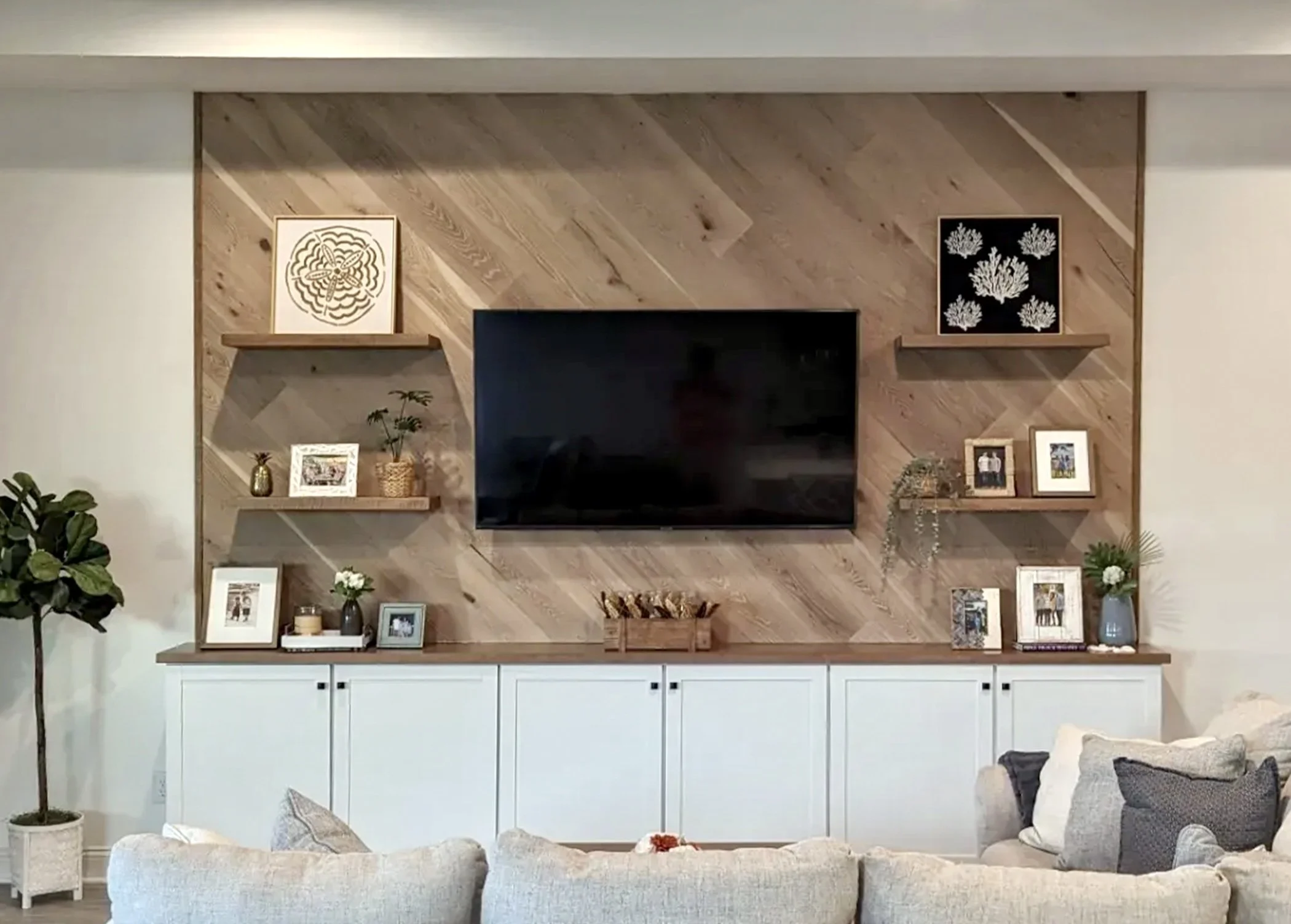 Living room with a wooden accent wall, a mounted flat-screen TV, and framed artwork. White cabinetry below the wall, decorated with framed photos, vases, and plants. A cream sofa with pillows in the foreground and a potted plant to the left.