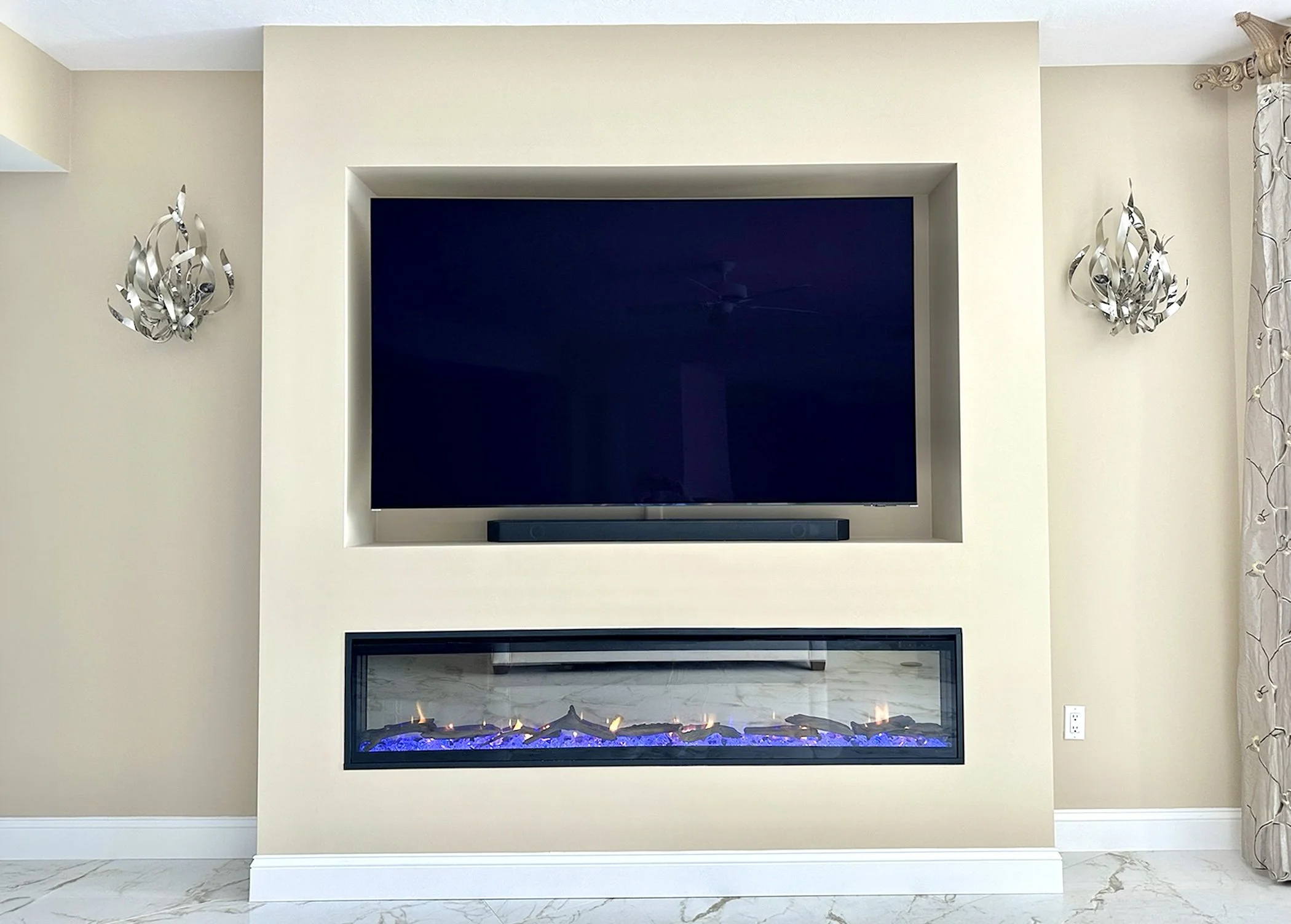 Modern living room with a wall-mounted flat-screen television above an electric fireplace, flanked by decorative chrome wall sconces, with beige walls and a marble floor.