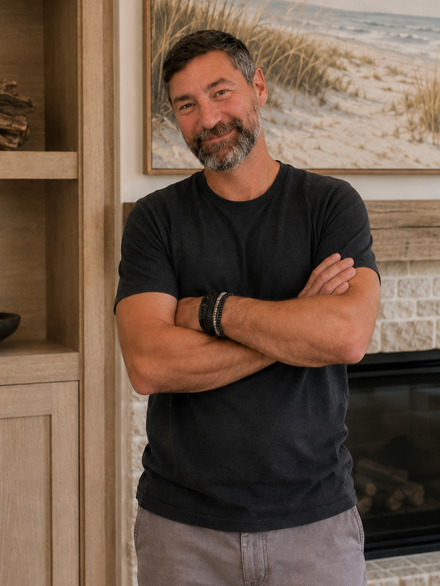 A man with a beard and mustache smiling, standing indoors with arms crossed, in front of a painting of a beach scene and a fireplace.