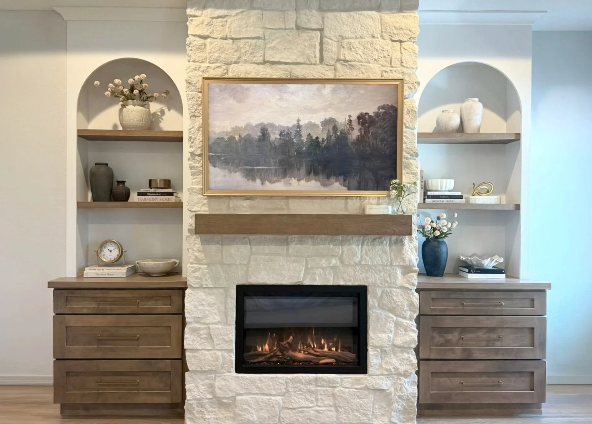 Living room fireplace with a stone surround, flanked by built-in shelves holding decorative vases, books, and plants, with a landscape painting above the mantel and a warm fire inside.