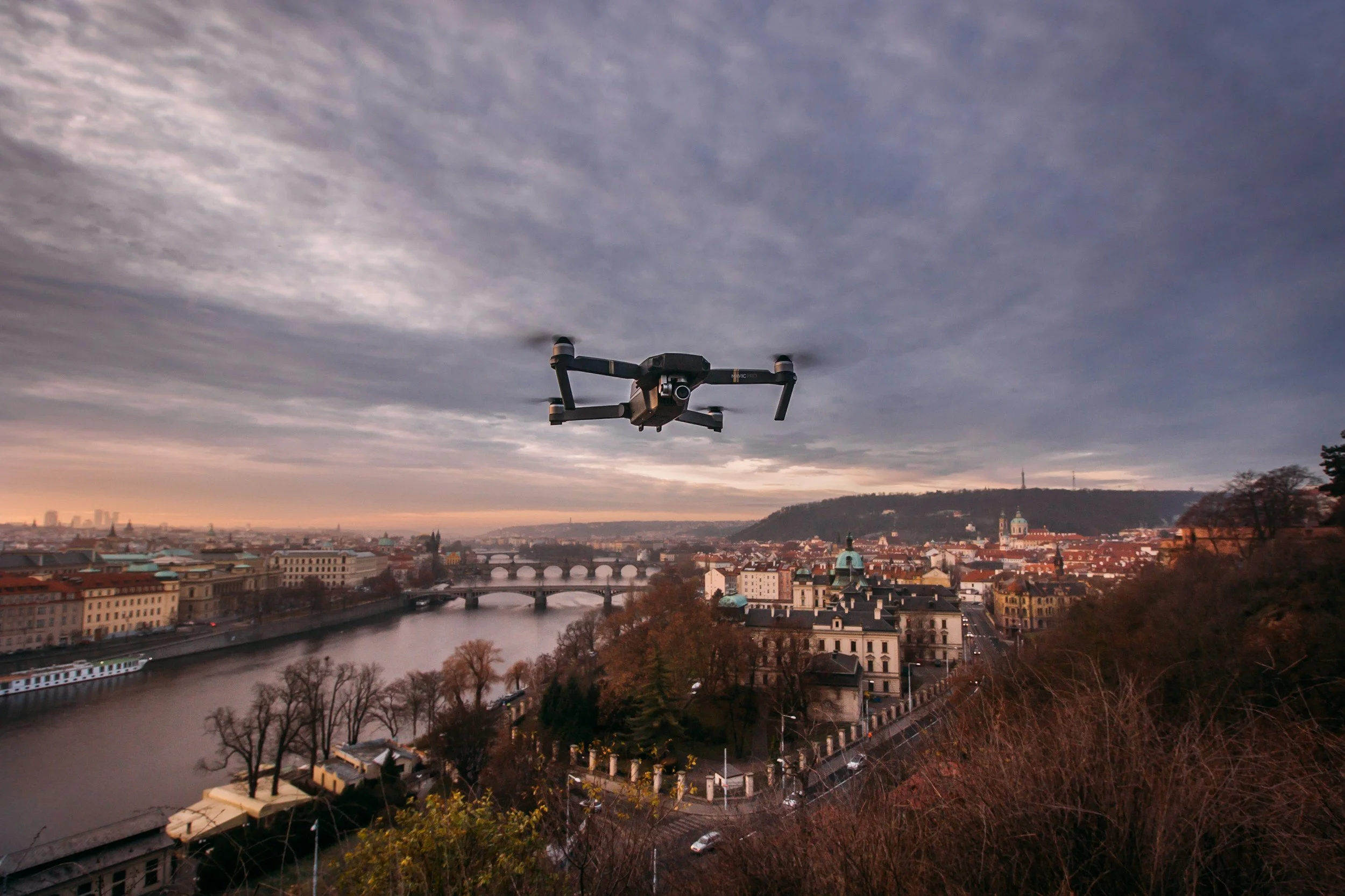 A drone flying over a cityscape with a river, bridges, and historic buildings during sunset, with a cloudy sky overhead.
