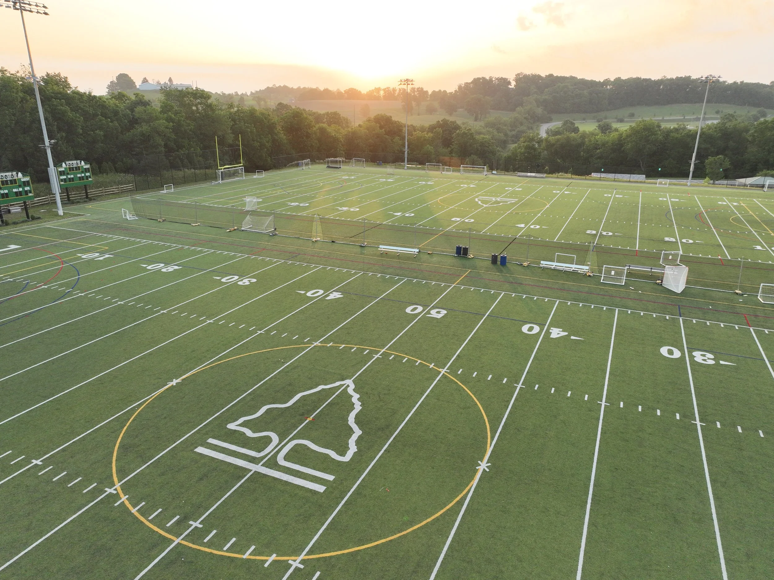 An outdoor football field with yard lines, goalposts, and soccer goals, surrounded by greenery and trees, with a sunset in the background.
