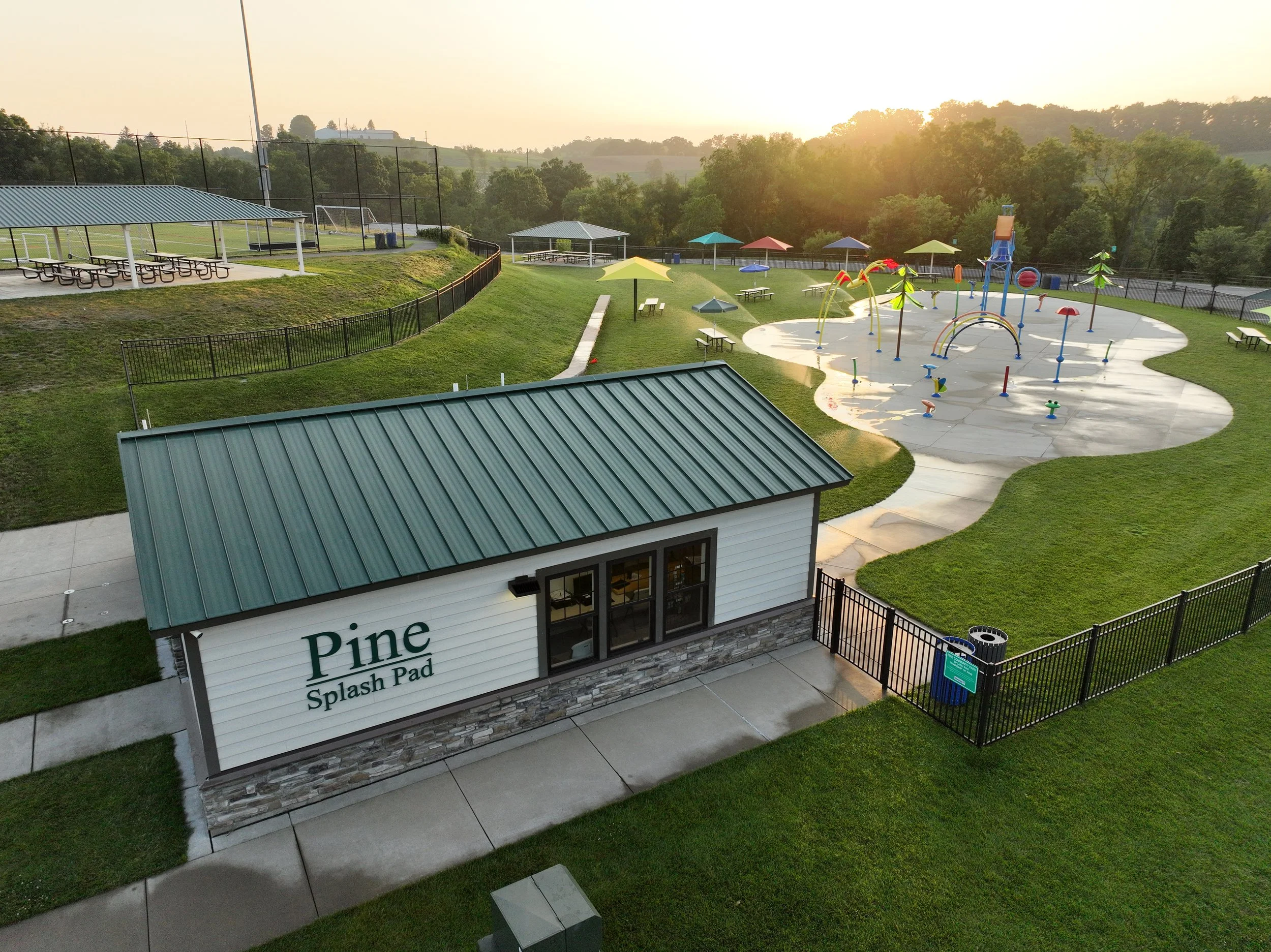 An outdoor splash pad at Pine Event Park with colorful umbrellas, water play structures, and a grassy area, set against a scenic background with trees and hills during sunset.
