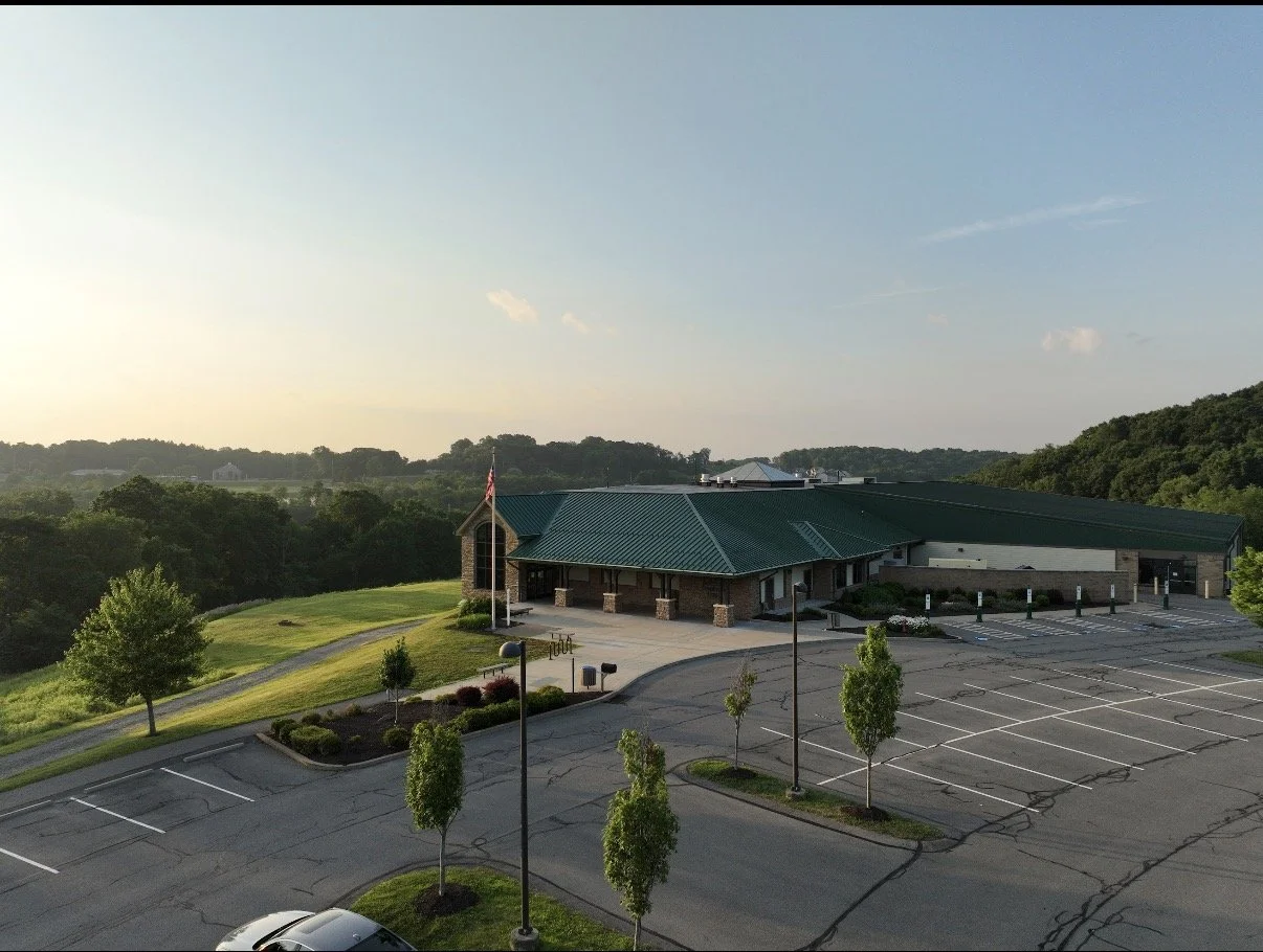 An empty parking lot in front of a building with a green metal roof, surrounded by trees and greenery, with a flag pole and scenic hills in the background during sunset.
