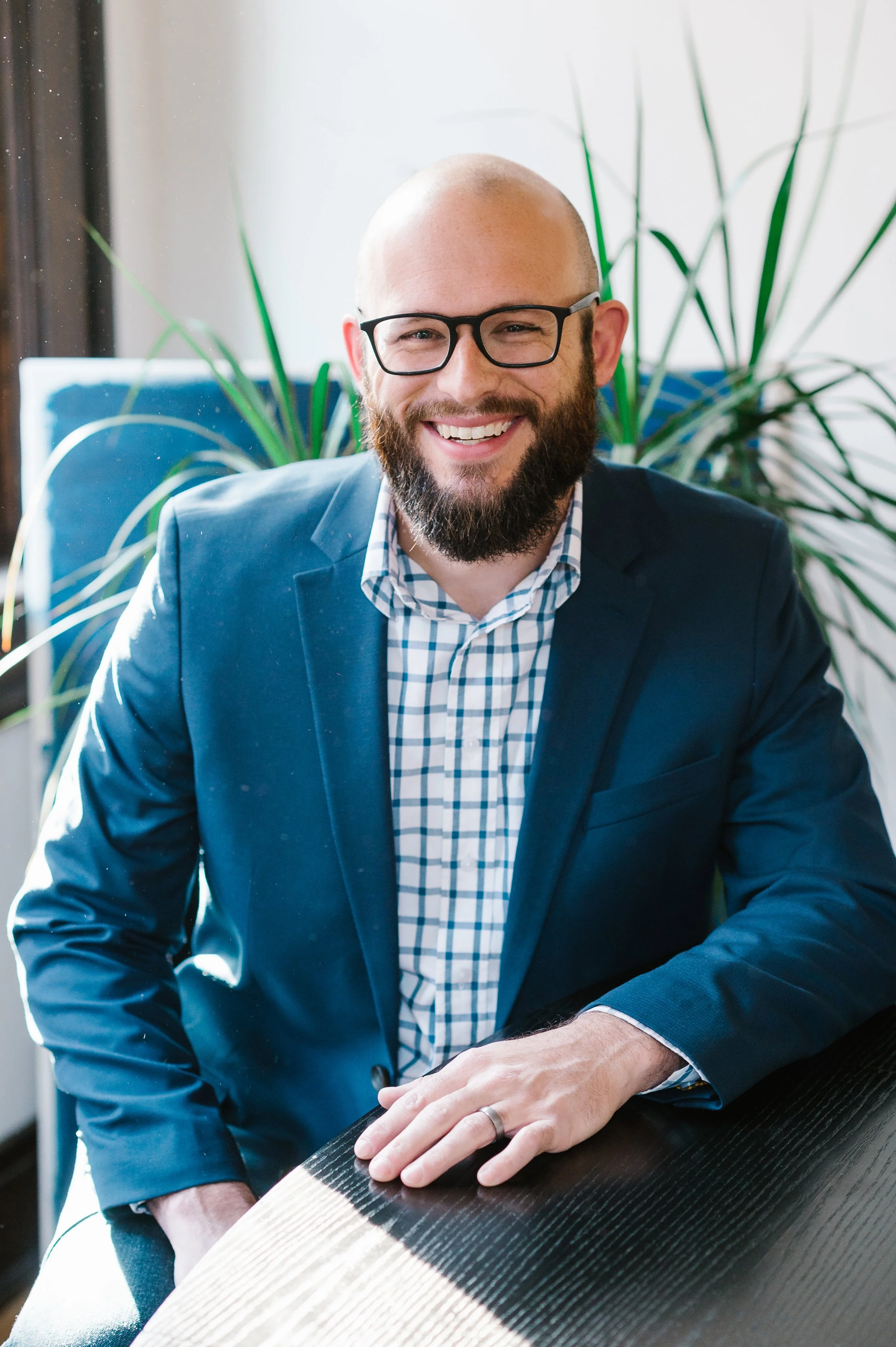A smiling man with a beard and glasses, wearing a blue suit jacket and checkered shirt, sitting at a table.