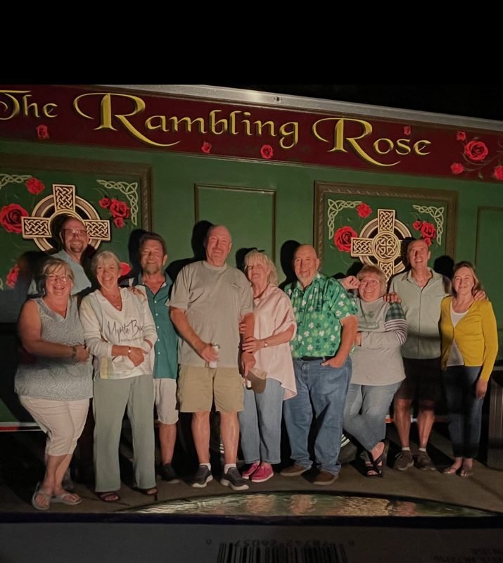 Group of nine people standing together in front of a green wall with Celtic crosses and roses at a pub called 'The Rambling Rose'.