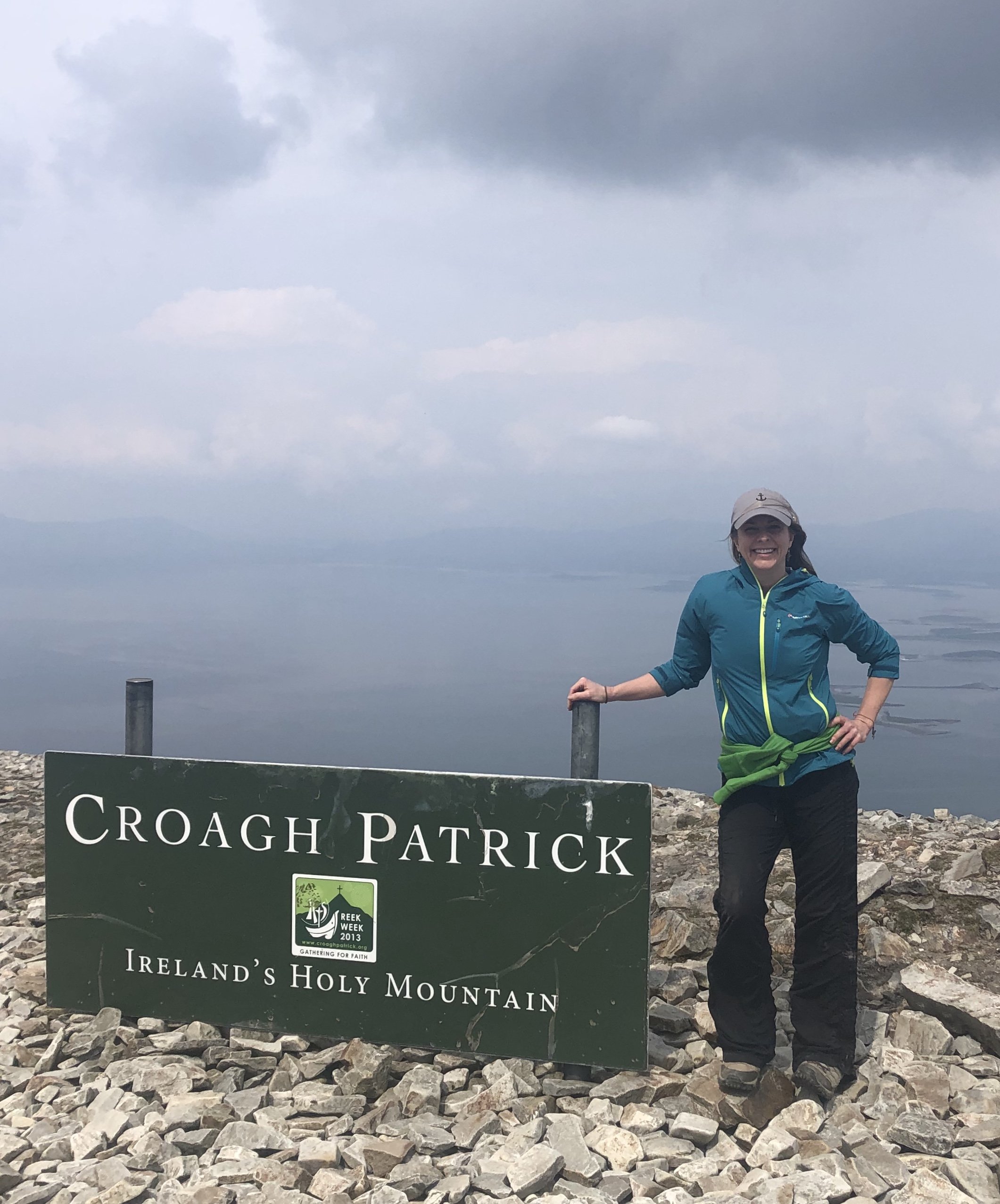 A smiling woman in outdoor clothing standing next to a large sign for Croagh Patrick, a mountain in Ireland, with a cloudy sky in the background.