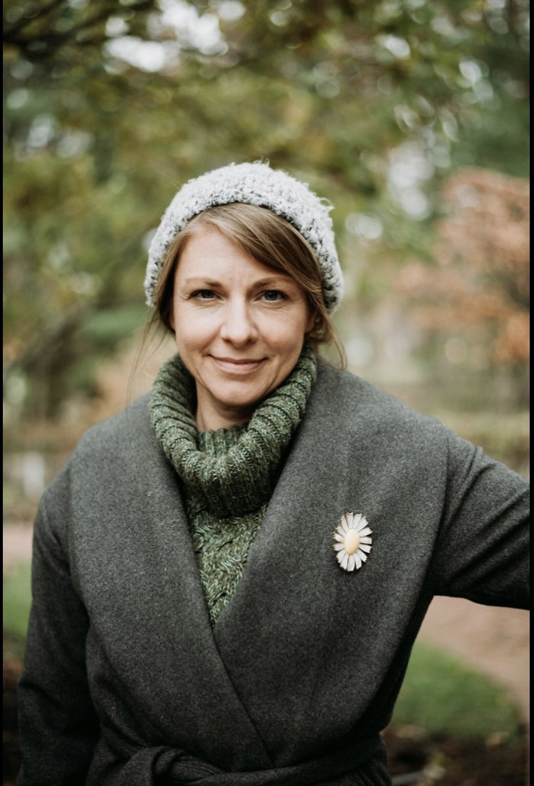 A woman outdoors in fall or winter attire, wearing a gray coat with a daisy-shaped brooch, a green sweater, and a knitted hat, with blurred autumn trees in the background.