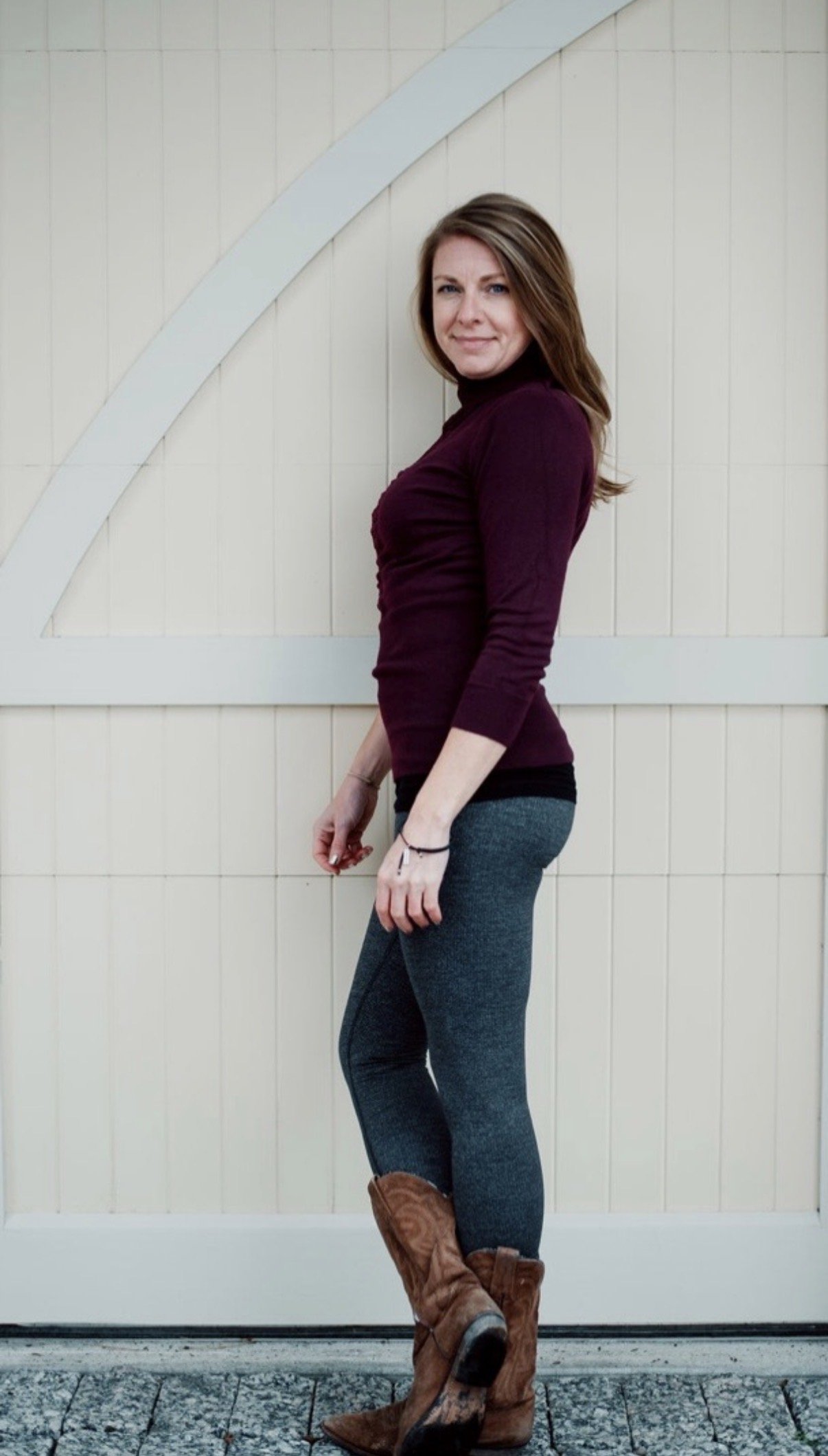 Woman with brown hair in a purple turtleneck, gray jeans, and brown cowboy boots standing against a beige wall with a diagonal white trim.