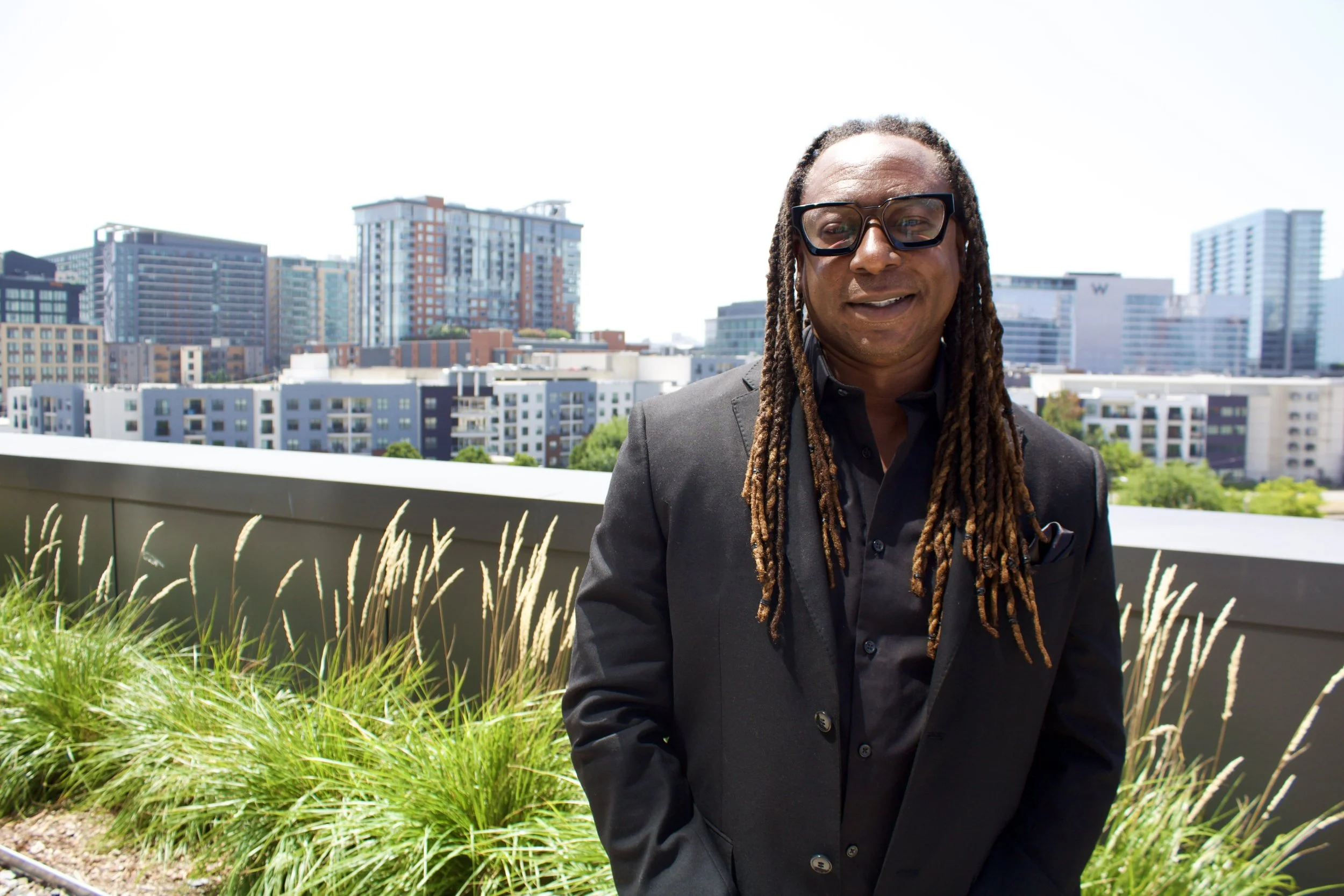 A man with long dreadlocks wearing glasses and a black suit standing outdoors on a balcony with a city skyline in the background.