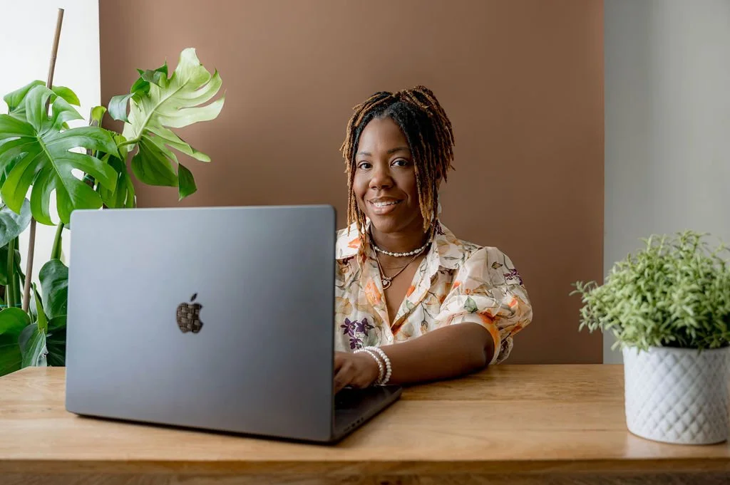 A woman with dreadlocks, smiling, sitting at a wooden desk with a silver Apple MacBook in front of her. She is wearing a floral blouse, pearl necklace, and bracelets. There are green plants on either side of her, one large monstera behind her and a smaller potted plant to her right, against a muted brown and gray background.