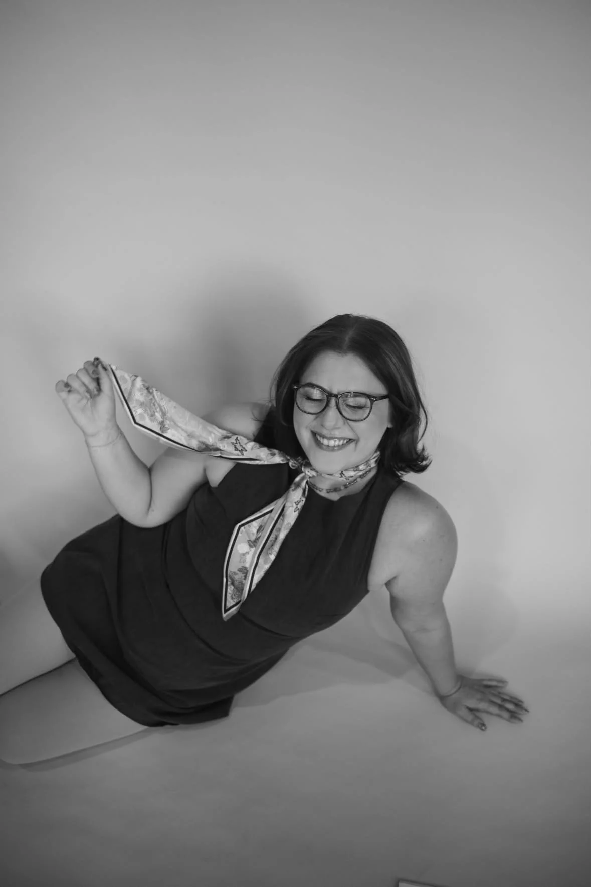 A woman with glasses and short hair, smiling and wearing a sleeveless dress, poses on the floor with one arm supporting her and one hand on the ground, holding a patterned scarf tied around her neck.