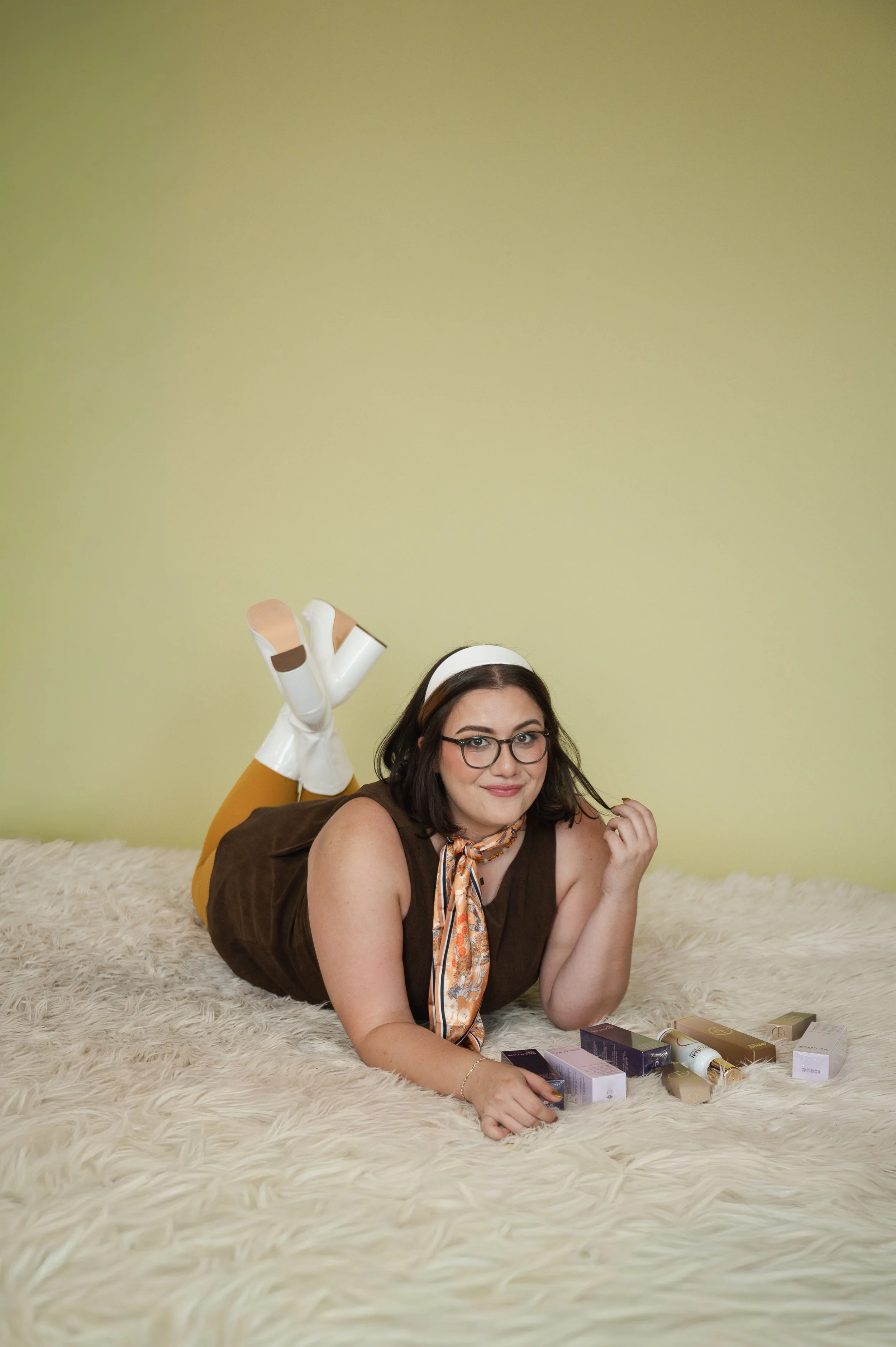 A woman with dark hair, glasses, wearing a brown sleeveless top and a silk scarf, lying on her stomach on a fluffy rug, smiling, with her chin resting on her hand, surrounded by makeup products.