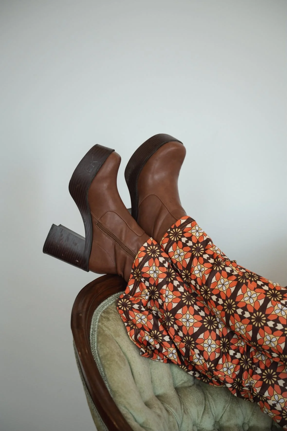Person wearing patterned pants and brown ankle boots, sitting on a vintage green upholstered chair.