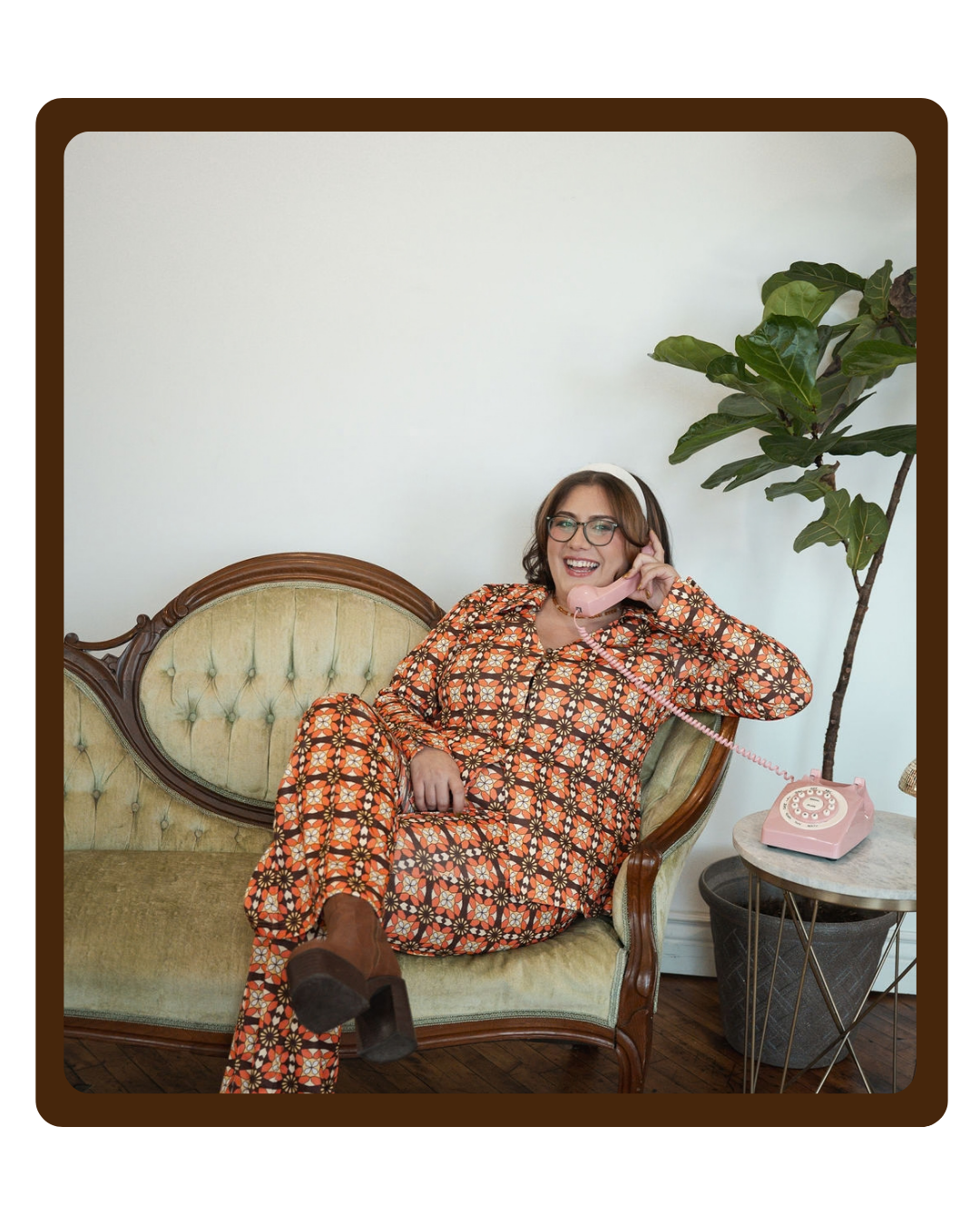 Woman in orange patterned jumpsuit sitting on a vintage green couch, smiling while talking on a pink rotary phone, with a large potted plant and a white wall in the background.