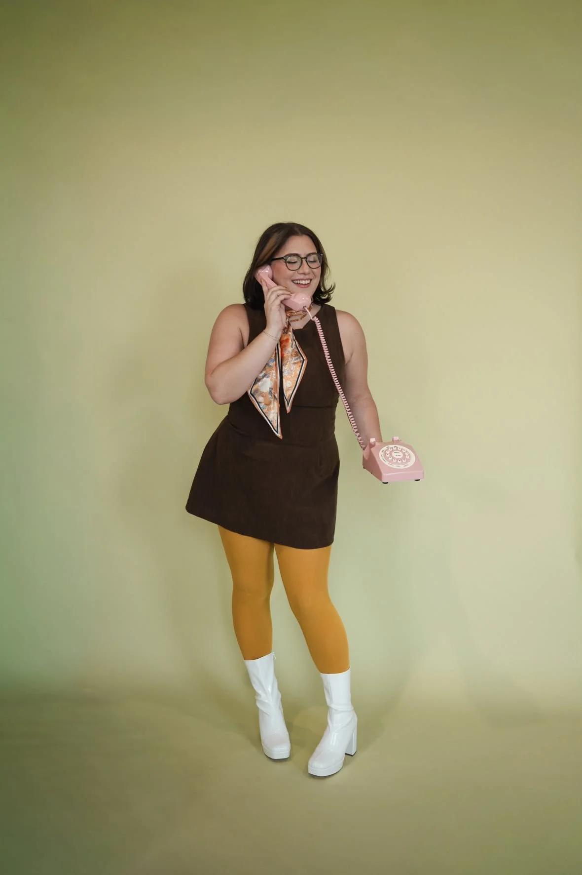 A woman with glasses, wearing a brown dress with a patterned scarf, yellow tights, and white platform boots, is smiling while talking on a pink vintage rotary telephone against a light green background.