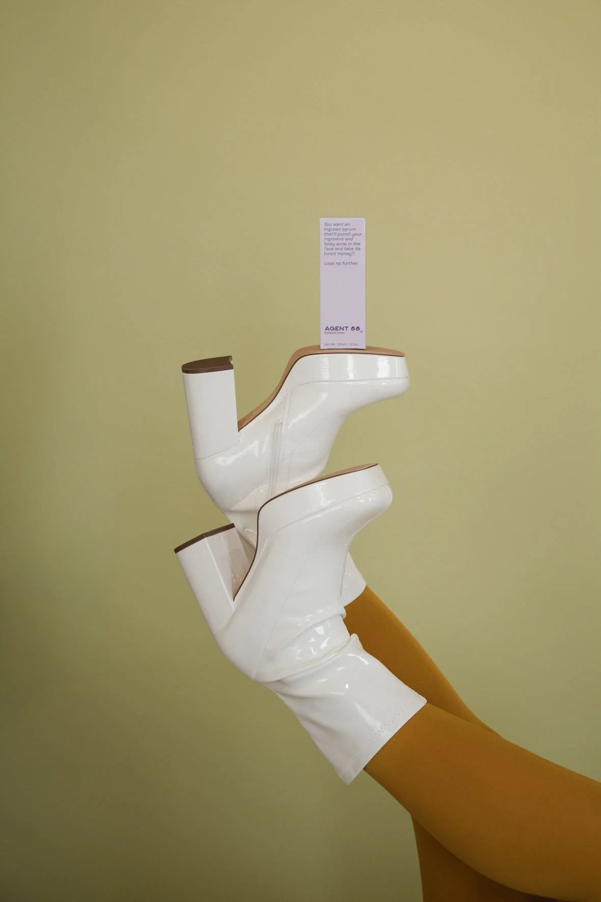 A person's legs wearing orange tights and white heeled boots with a boxed product balanced on the sole of the boot.