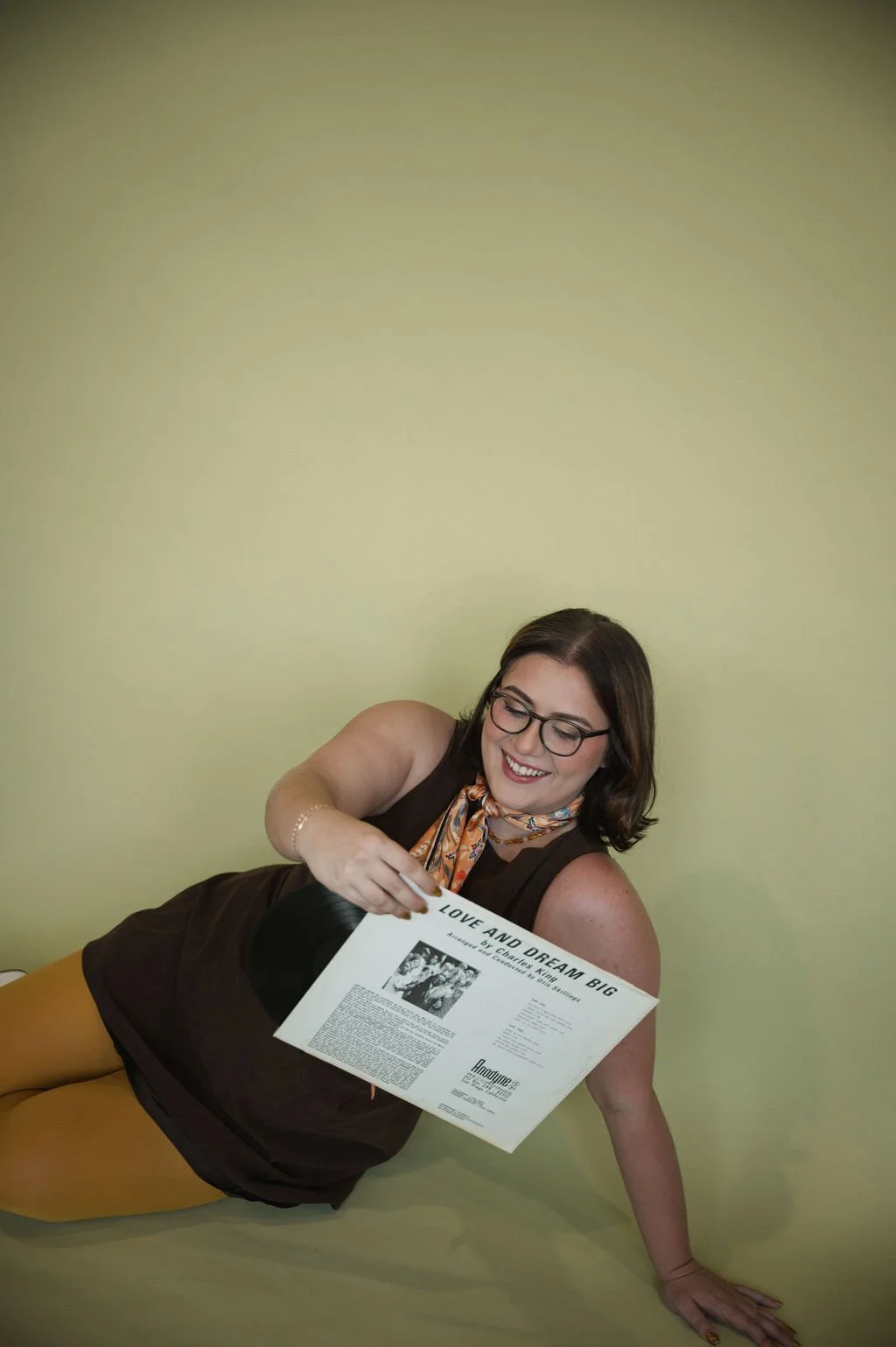 Woman with glasses lying on the floor against a plain wall, smiling, holding a vinyl record.