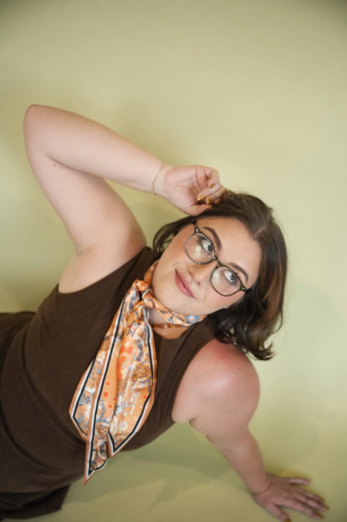 A woman with glasses and a scarf around her neck lying on the floor against a yellow wall, with her arm resting behind her head and smiling while looking slightly to the side.
