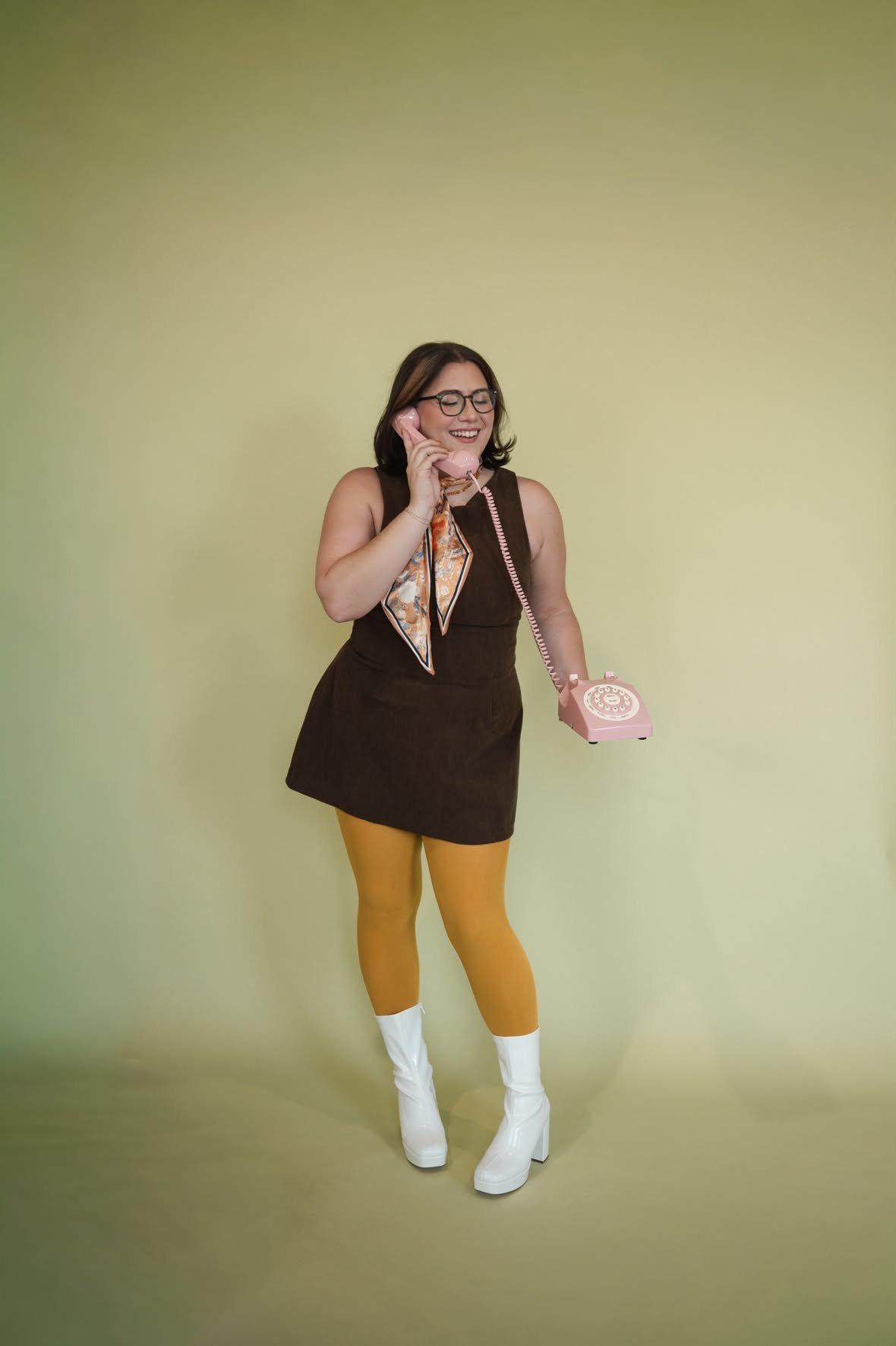A woman smiling and talking on a pink rotary phone, standing against a light green background. She is wearing a sleeveless dark brown dress, yellow tights, white boots, glasses, and a patterned scarf.