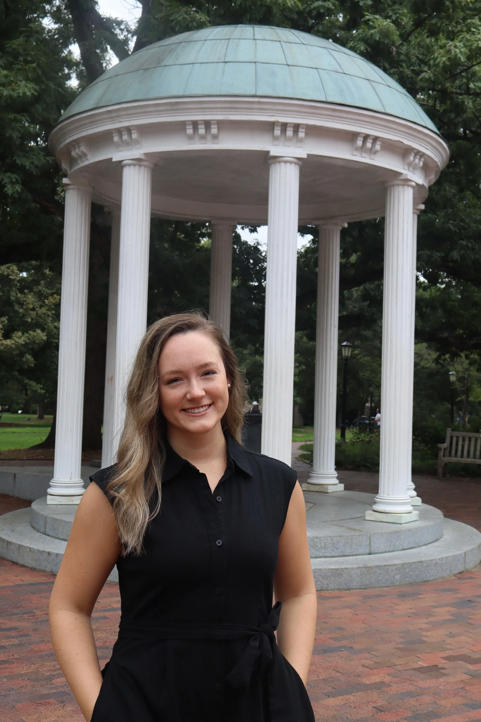 Young woman with long, styled hair smiling and standing outdoors in front of a classical white pavilion with columns and a domed roof, surrounded by trees and park benches.