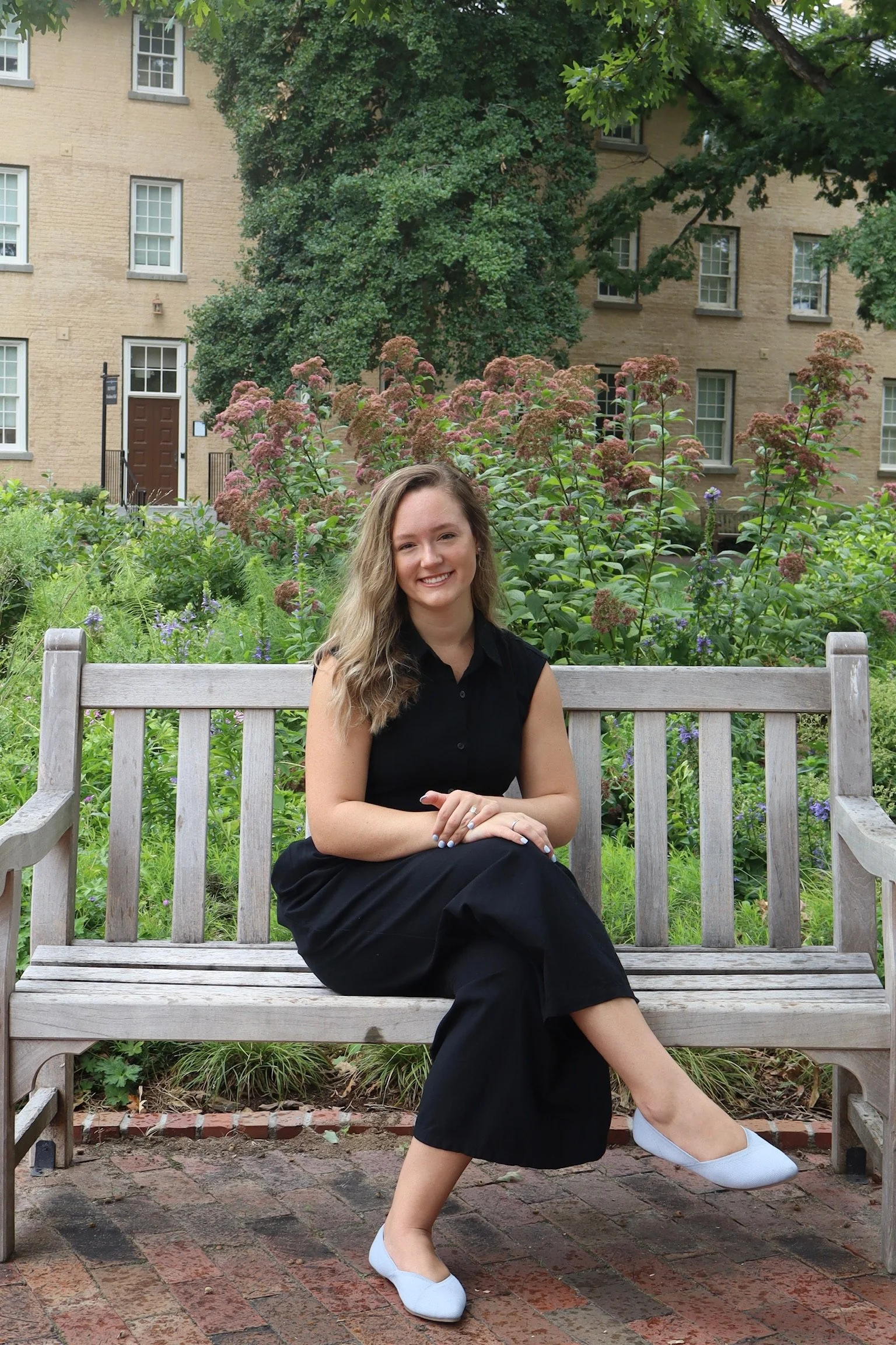 A woman sitting on a wooden bench outdoors surrounded by green plants and flowers, wearing a black sleeveless dress and white shoes, smiling at the camera, with a brick walkway and beige building in the background.