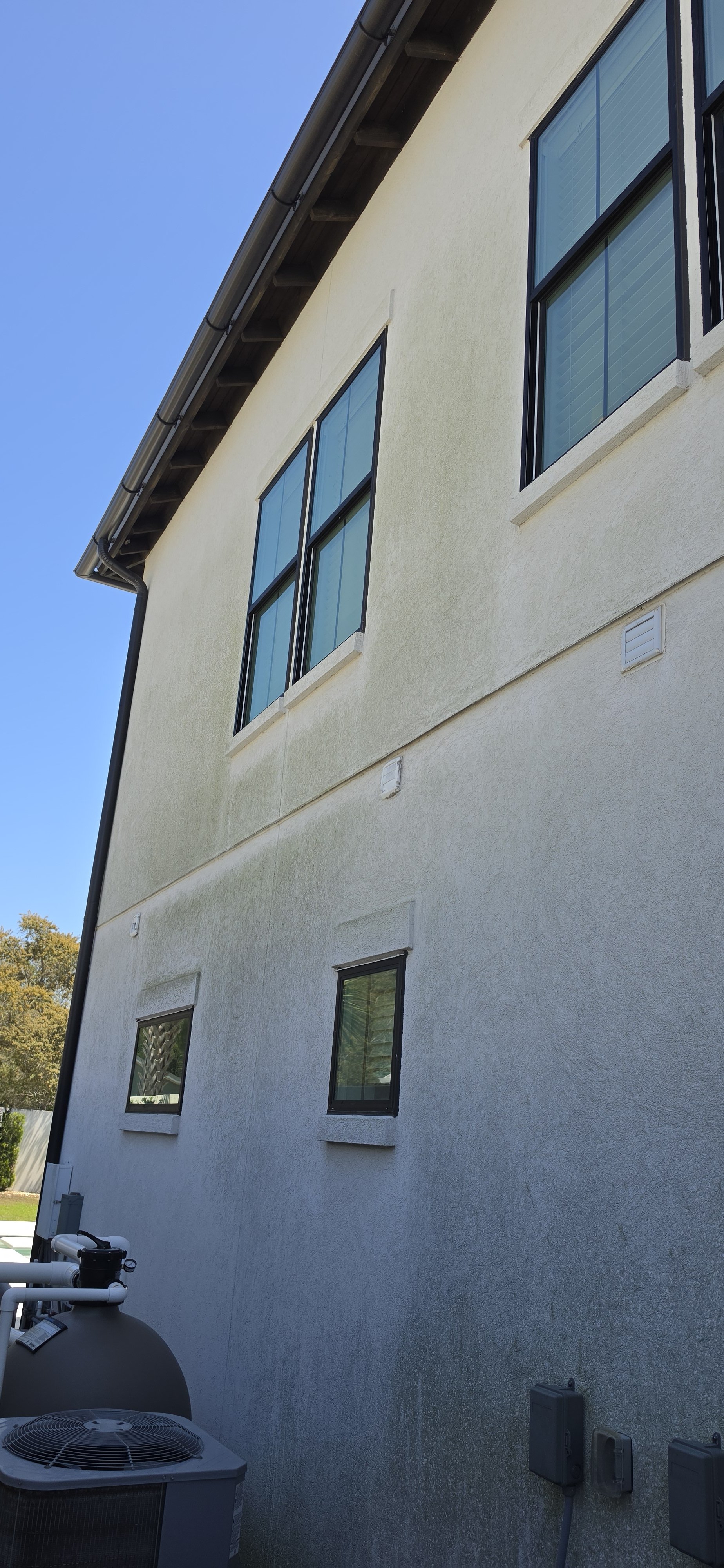 BEFORE PICTURE-Side view of a white multi-story residential building with black window frames, visible outdoor HVAC units, and a clear blue sky with mold on exterior of home