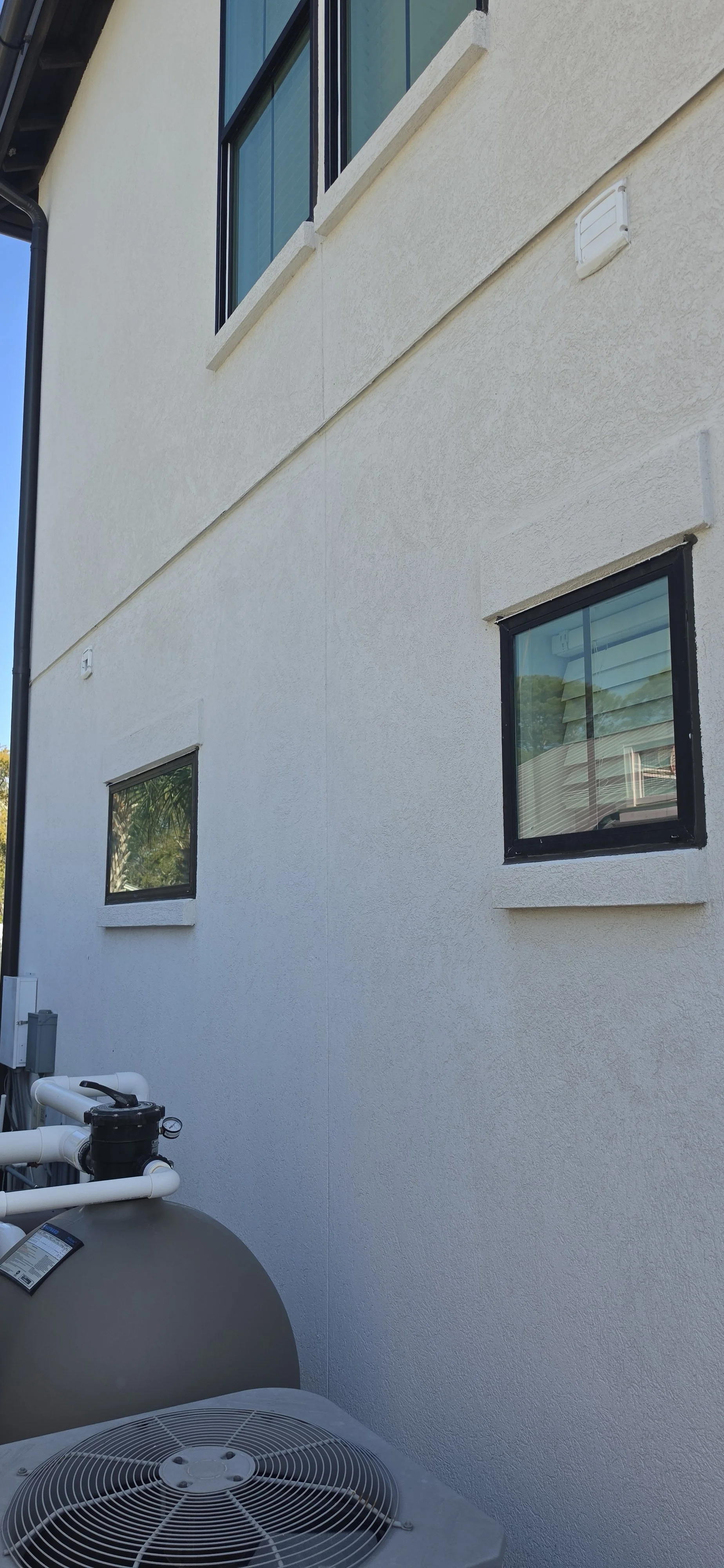 AFTER PICTURE-View of the side of a white stucco building with three windows, a rain gutter, and an external air conditioning unit after cleaning