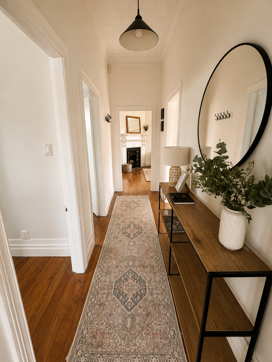  hallway with wooden floor, beige patterned runner rug, white walls, black-framed console table with decorative items, large round mirror, table lamp, plants, and a view into other rooms with fireplaces in the background 