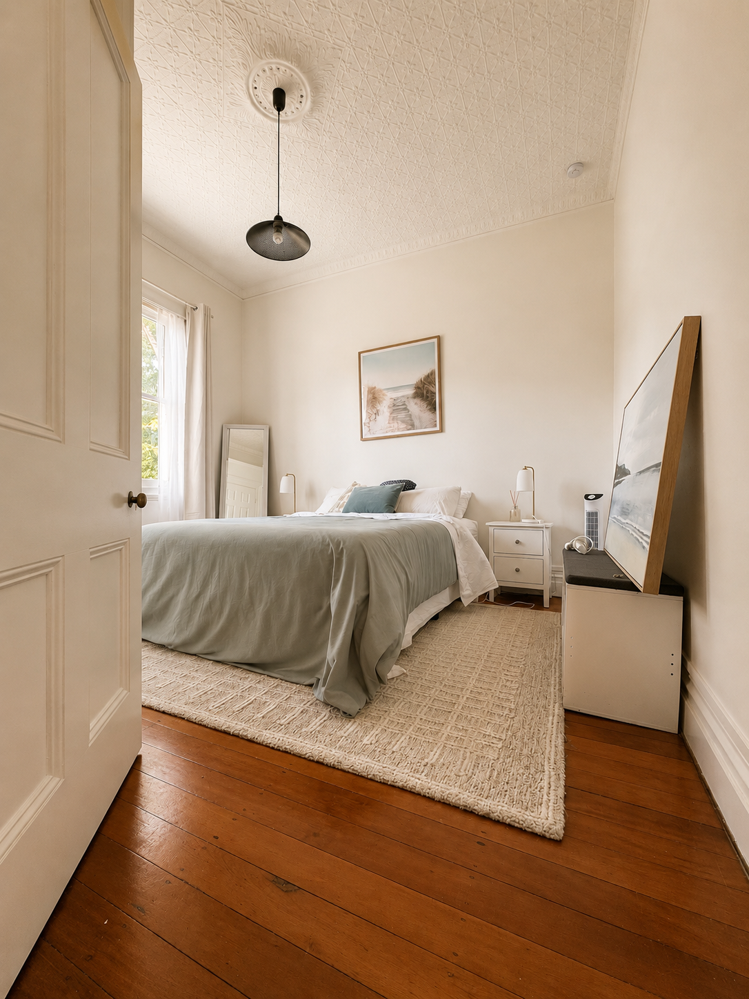 A cozy bedroom with white walls, a bed with gray bedding, a white nightstand, a framed picture of a beach scene, a mirror, a floor lamp, a TV on a cabinet, and hardwood flooring.