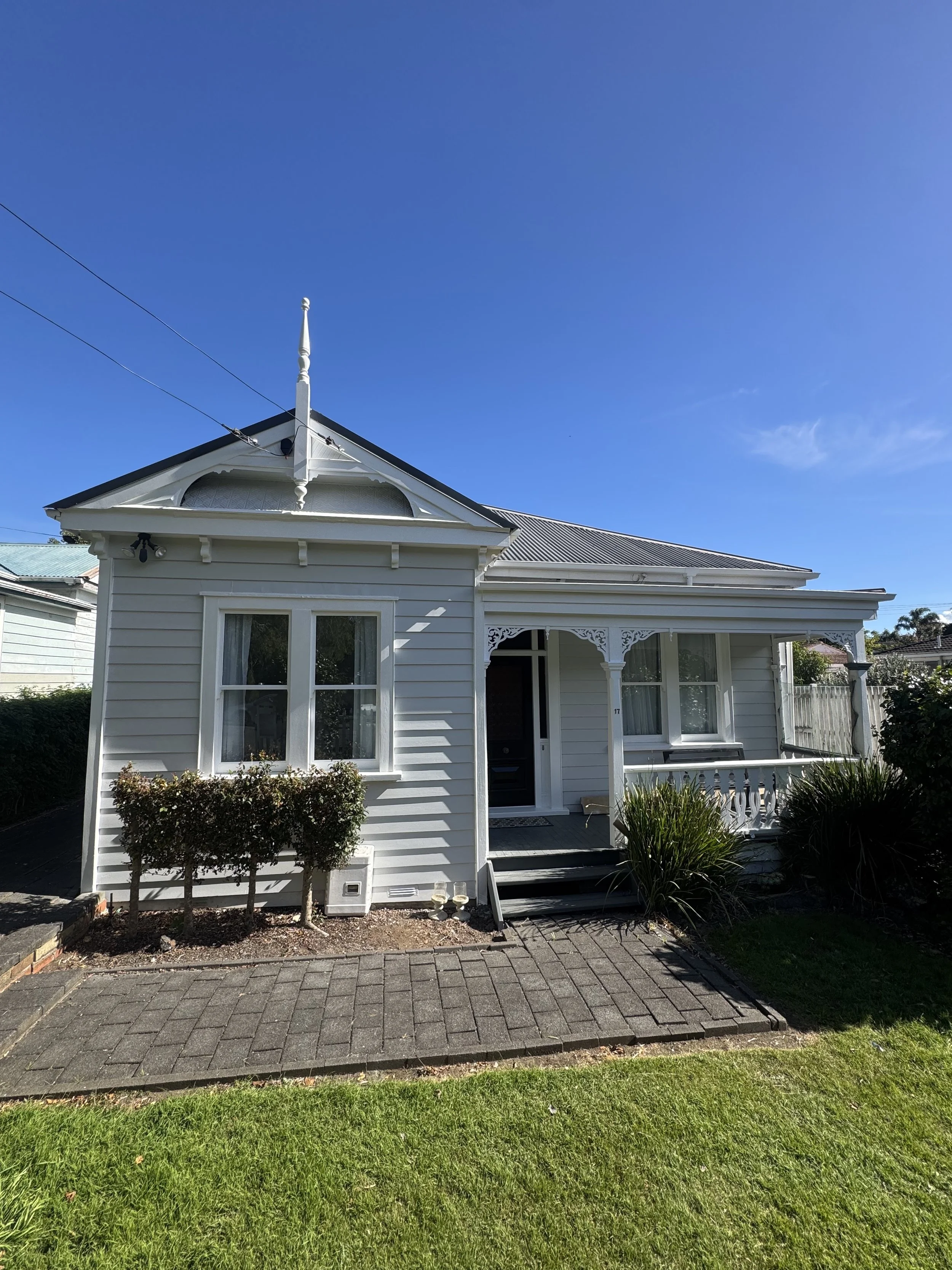 Front view of a white, Victorian-style house with a porch, surrounded by a small garden with shrubs, on a paved driveway, under a clear blue sky.