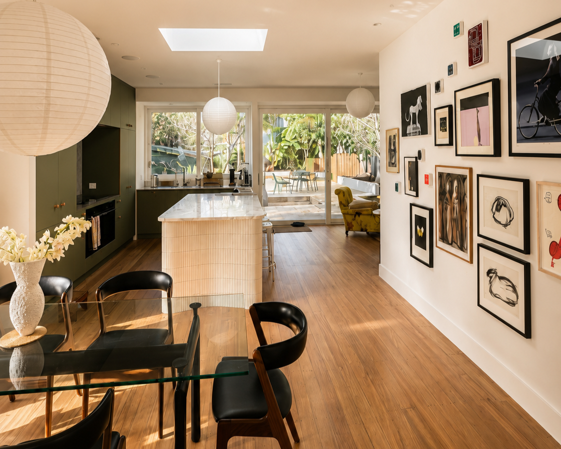 Interior view of a modern open-concept kitchen and dining area with wood flooring, green cabinetry, a glass dining table with black chairs, a white marble kitchen island, and large sliding glass doors leading to an outdoor patio with greenery, artwork, and a yellow armchair.