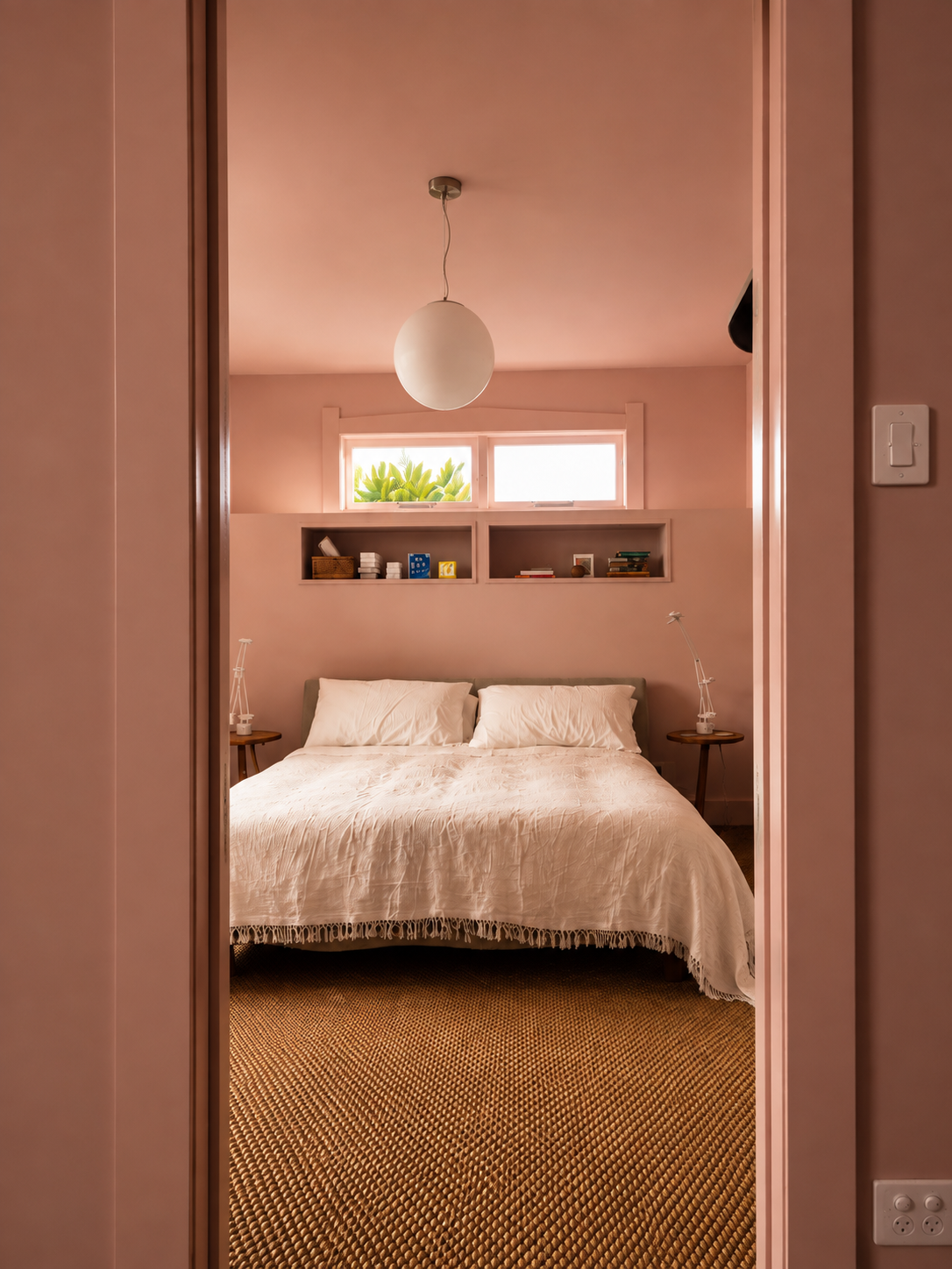 View of a bedroom through a doorway with pink walls, featuring a bed with white bedding, two wooden side tables with white lamps, a small window above the bed, and built-in shelves with books and decor.