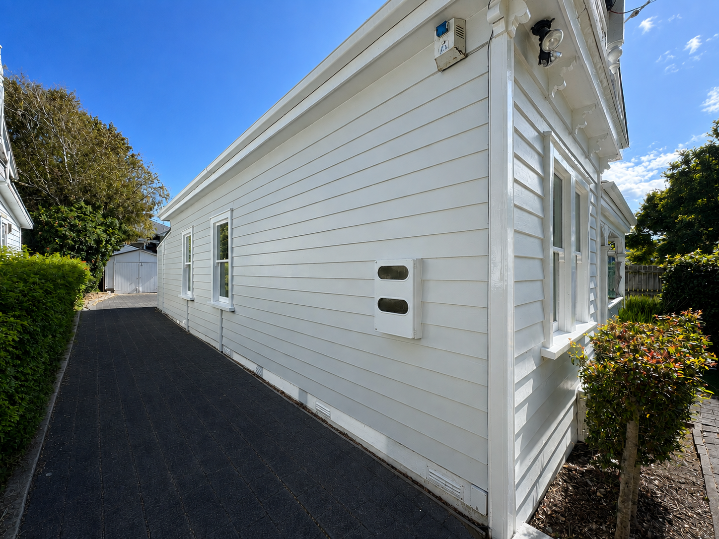 Side view of a white house with horizontal siding, three windows, and a black pathway. Trees and bushes are visible around the house under a bright blue sky.