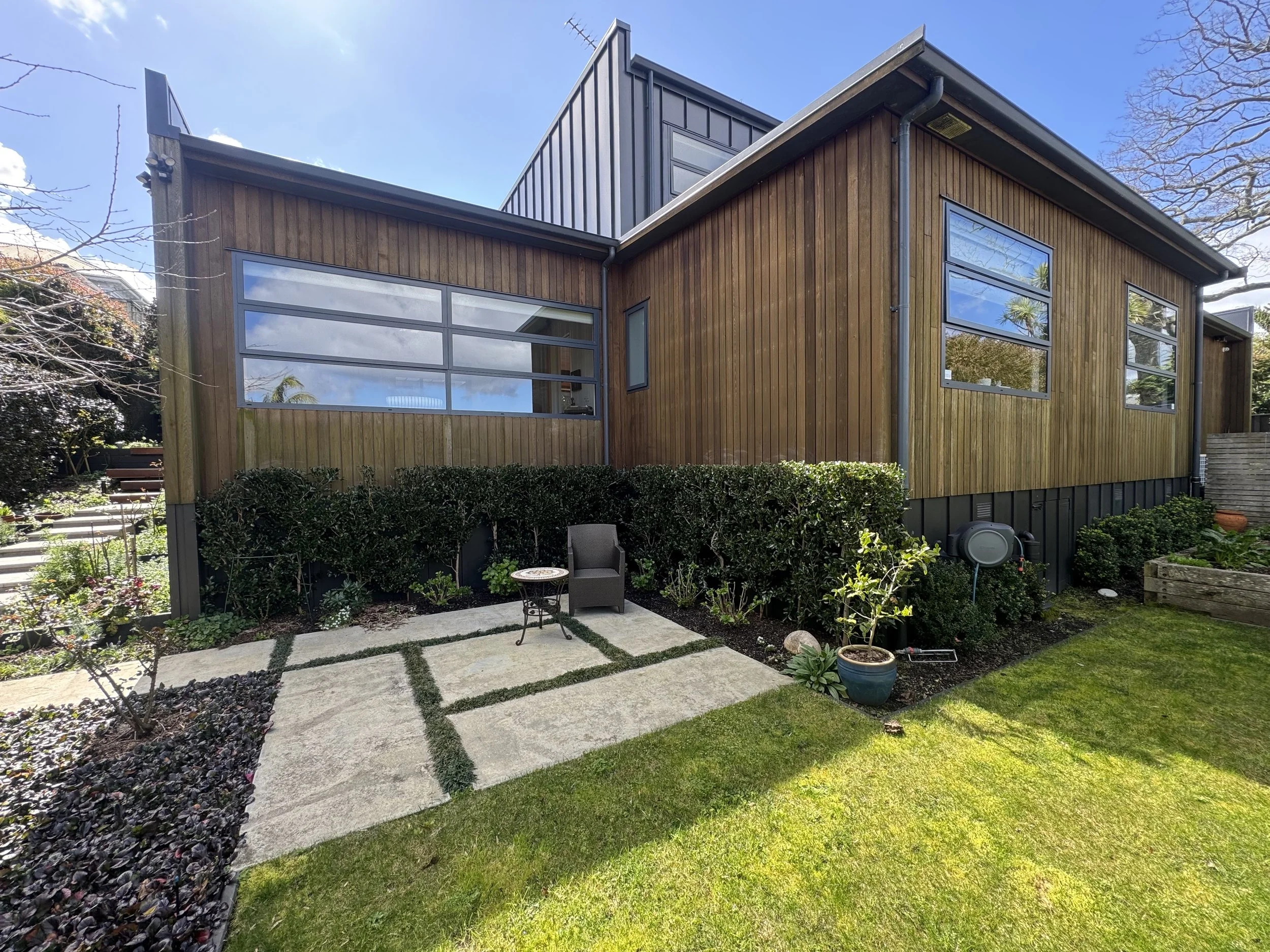 A modern house with wooden siding and large glass windows, surrounded by a manicured garden with concrete stepping stones, outdoor furniture, and lush green grass.