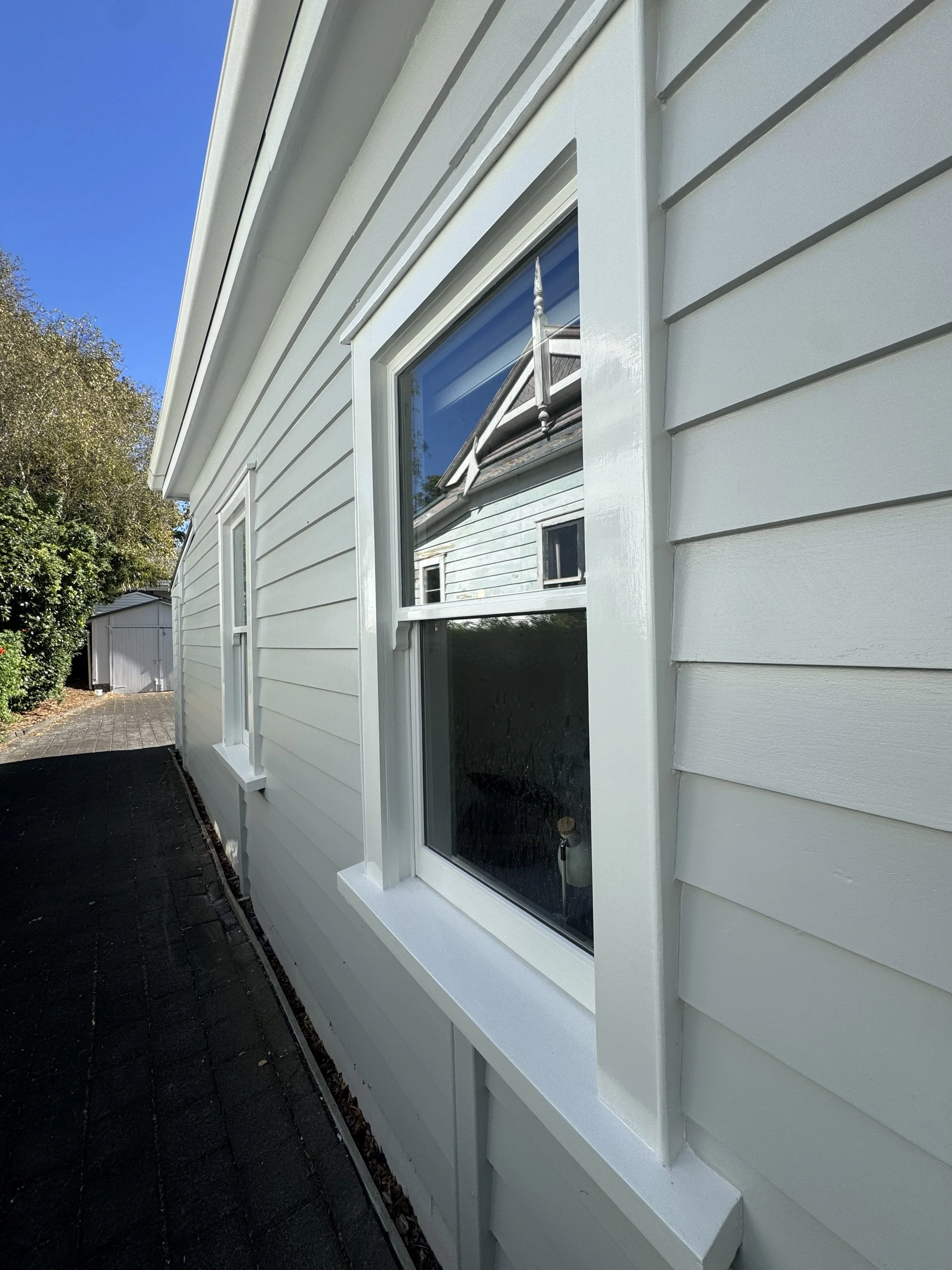 Close-up view of a house's exterior wall showing a window with a small metal tool leaning against the window frame, and reflecting the blue sky and nearby structures.