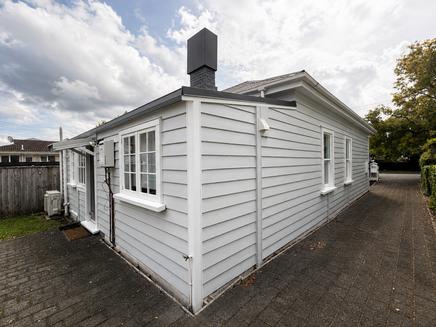 White house with horizontal siding, multiple windows, a paved backyard with a wooden fence and trees, cloudy sky.