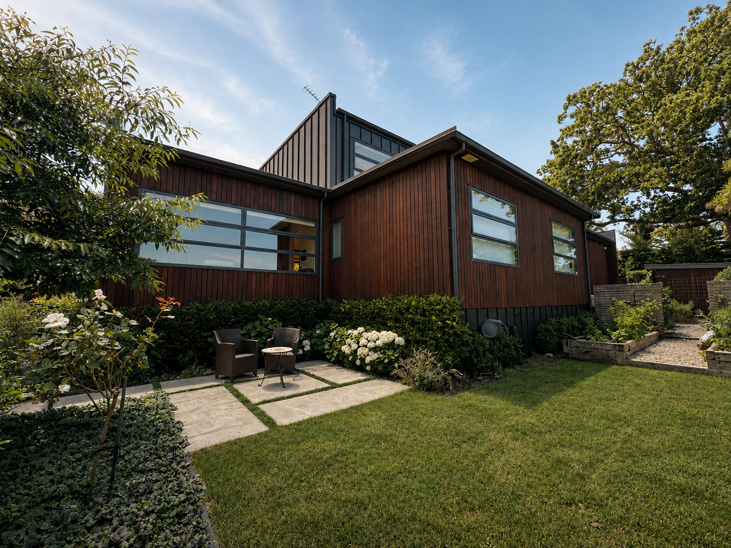 Modern house with wooden siding and large windows, surrounded by a well-maintained garden with seating area, bushes, flowers, and trees, under a partly cloudy sky.