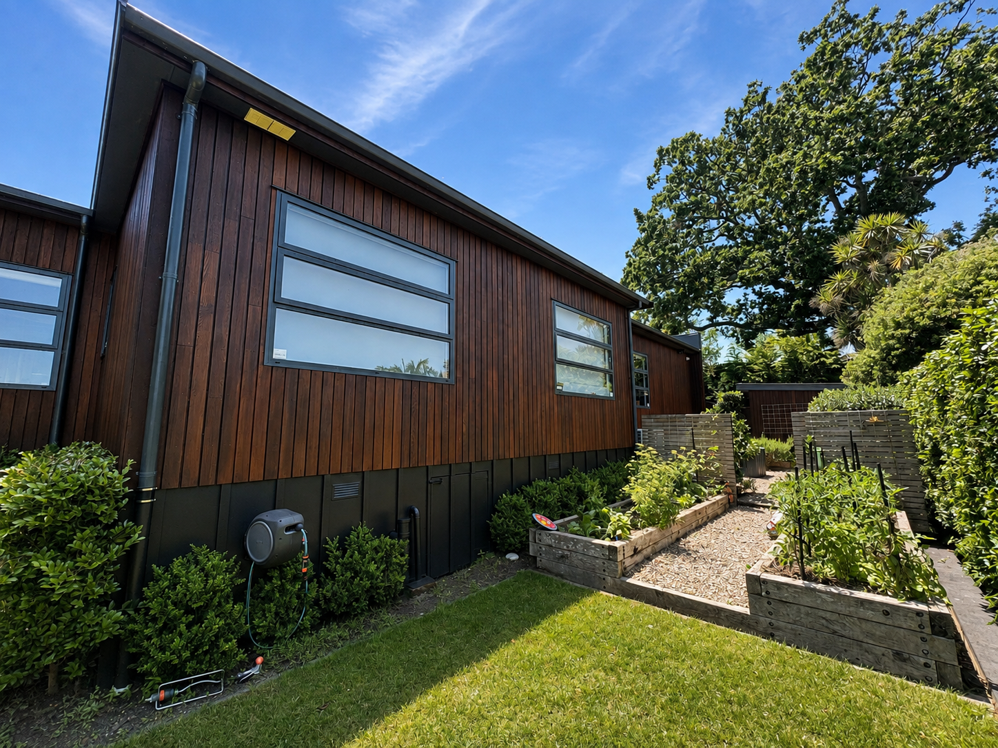 A modern house with wooden siding, large windows, and a backyard garden with raised vegetable beds, surrounded by lush greenery and a clear blue sky.