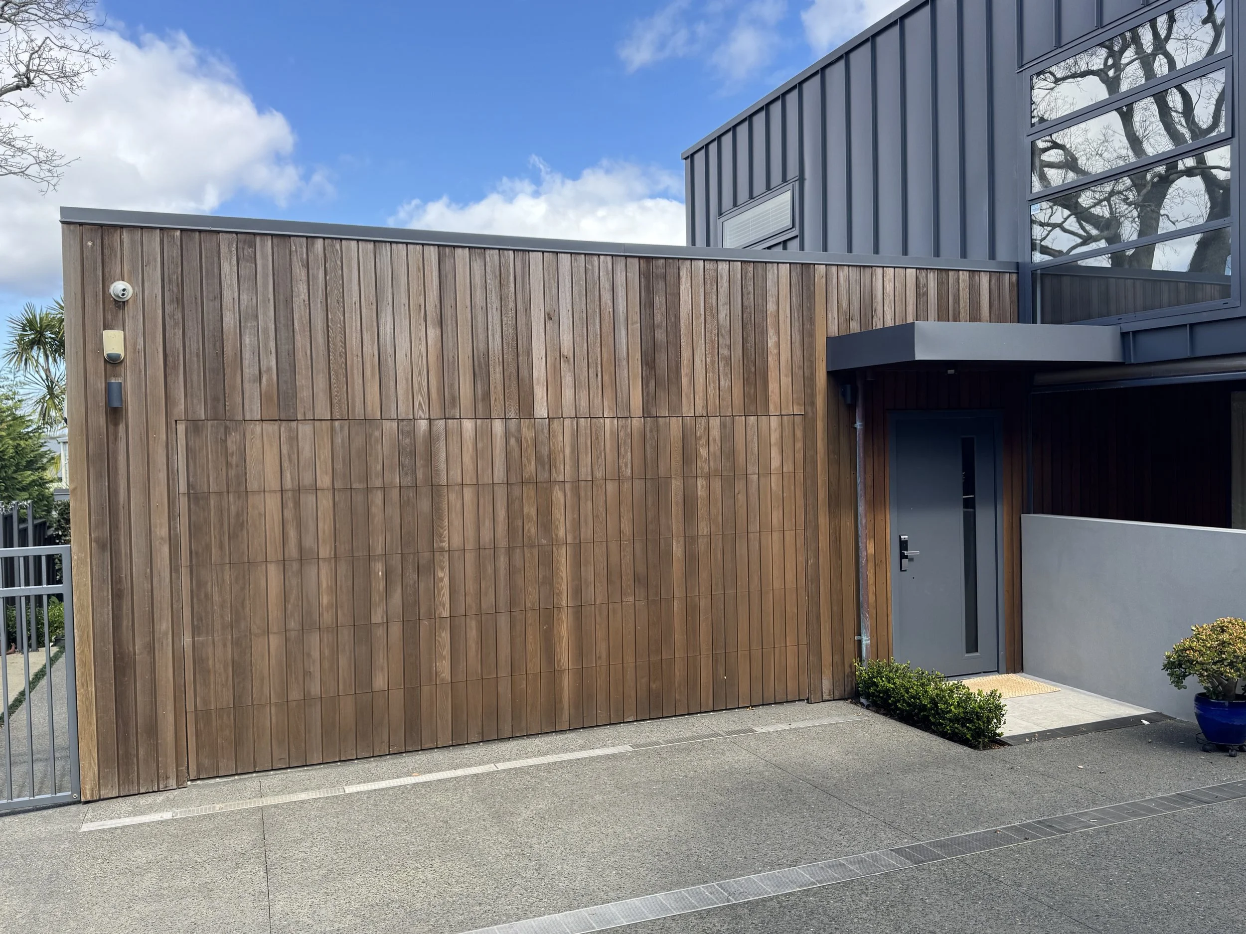 Modern building with wooden and metal siding, large glass windows, and a gray entrance door, surrounded by small bushes and a blue pot, under a partly cloudy sky.
